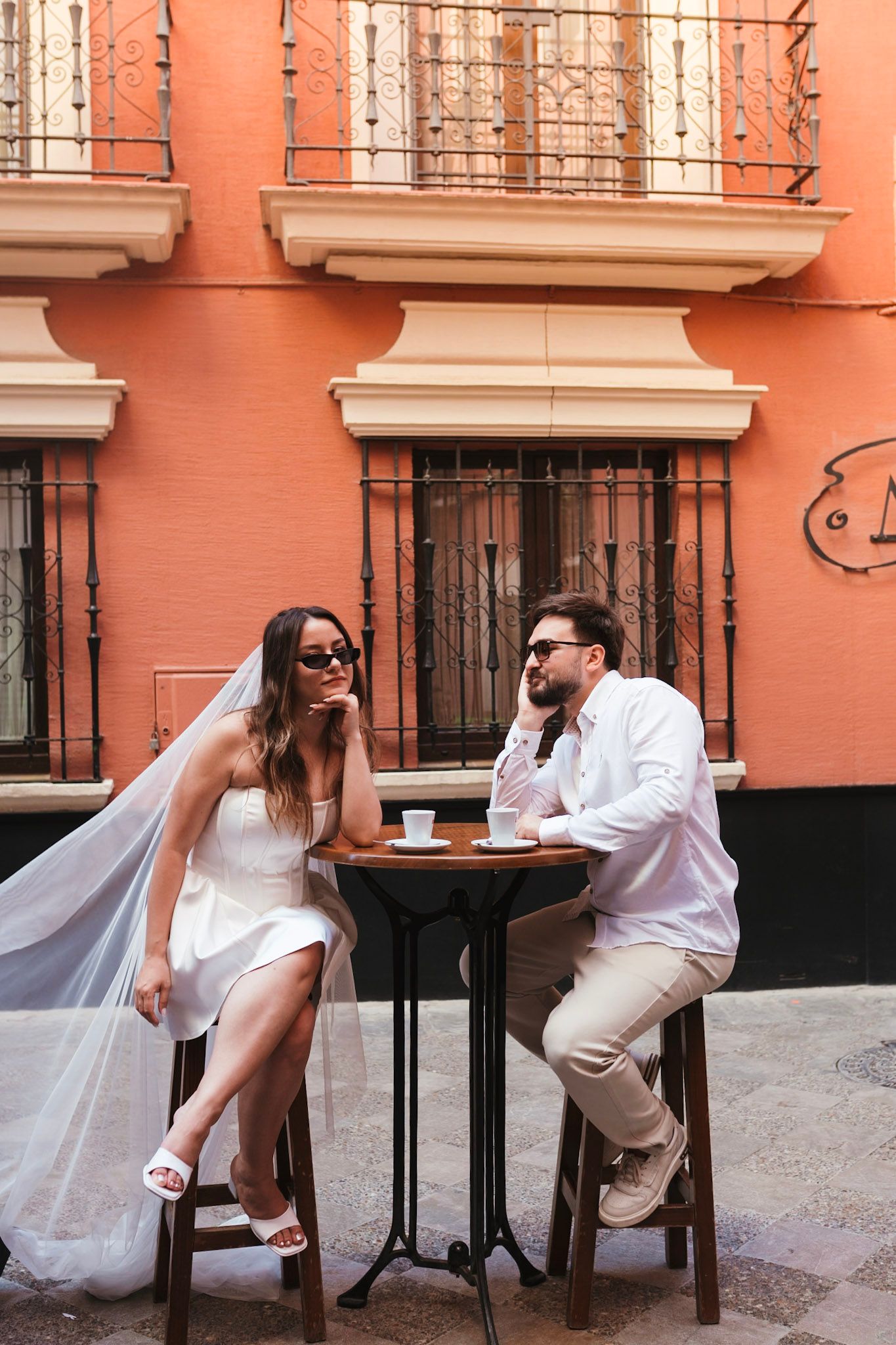 Bride in white dress and groom in white shirt sit at outdoor cafe table in front of a coral-colored building in Seville.
