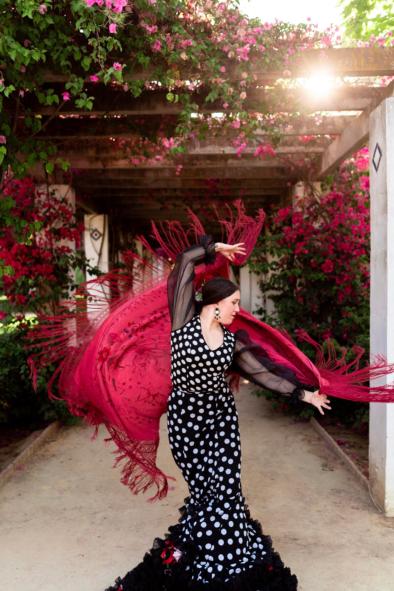 Flamenco dancer in polka dot dress swirling red fringed shawl among blooming pink flowers.