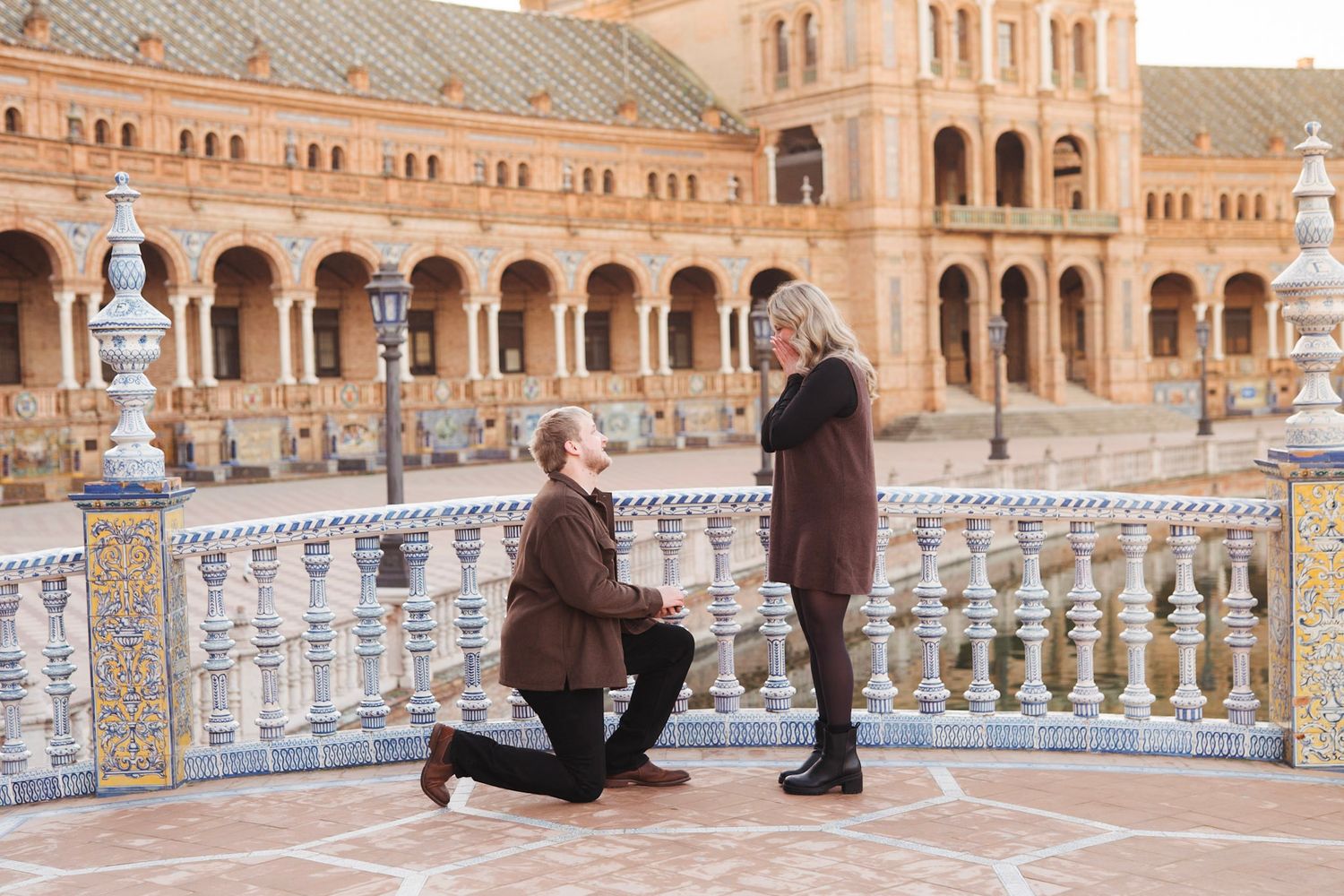 Man on one knee proposing to woman at ornate Plaza de Espana in Seville, Spain.