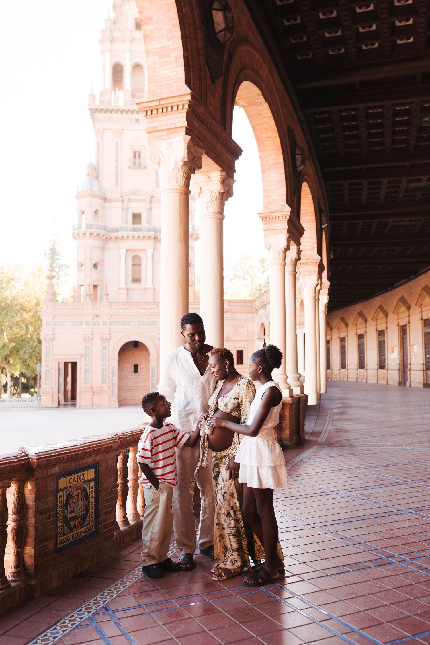 A joyful Black family of four stands together under ornate arches at Plaza de España, Seville, Spain.