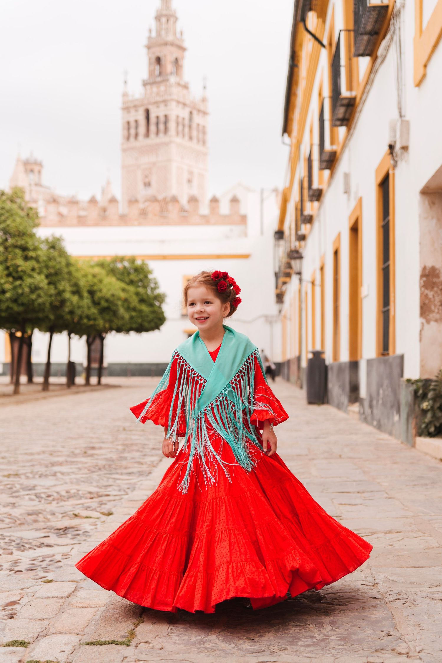 Young girl in red and turquoise flamenco dress twirls on cobblestone street in Seville.