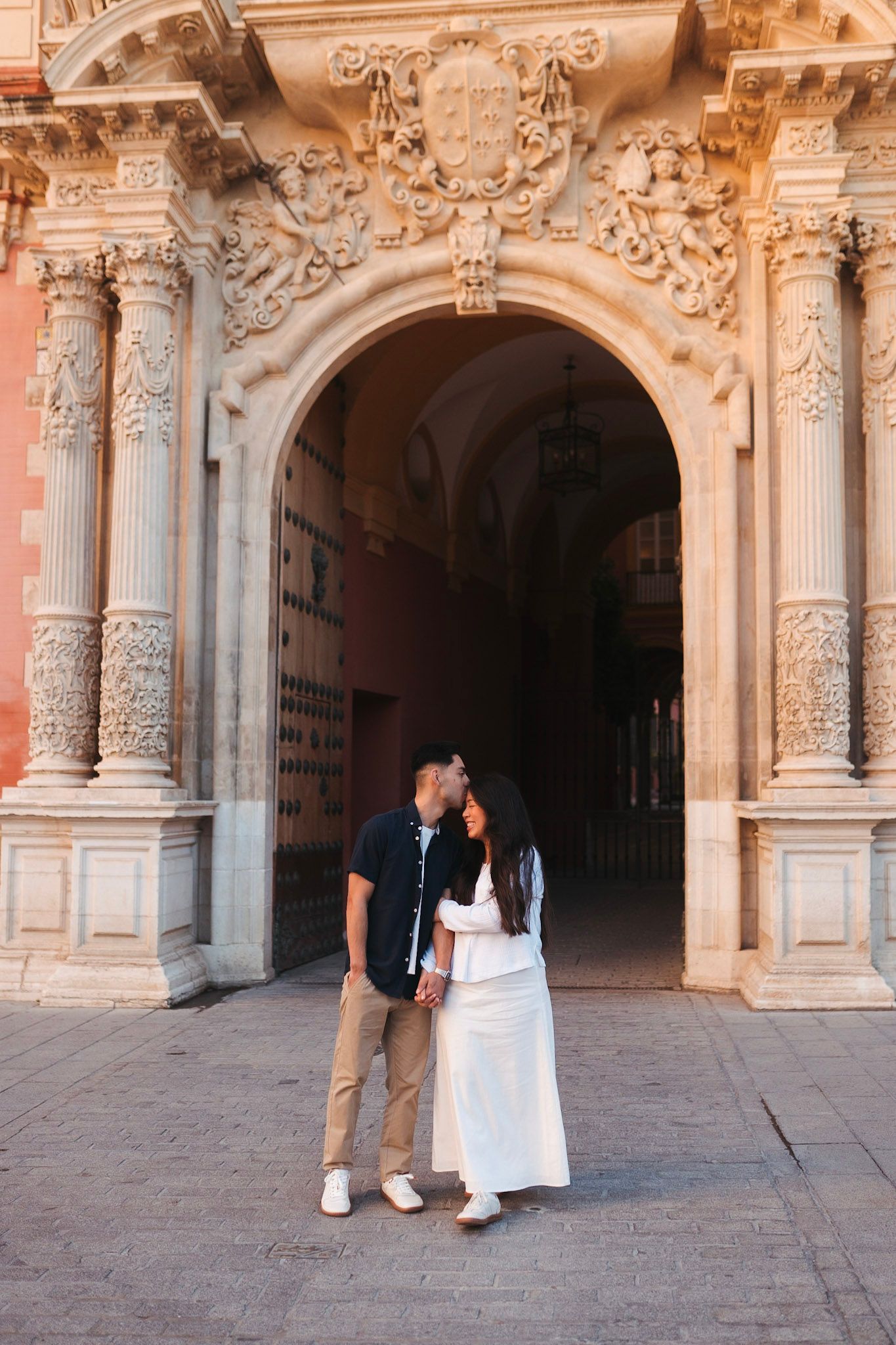Couple sharing a kiss under an ornate baroque stone archway in Seville, Spain.