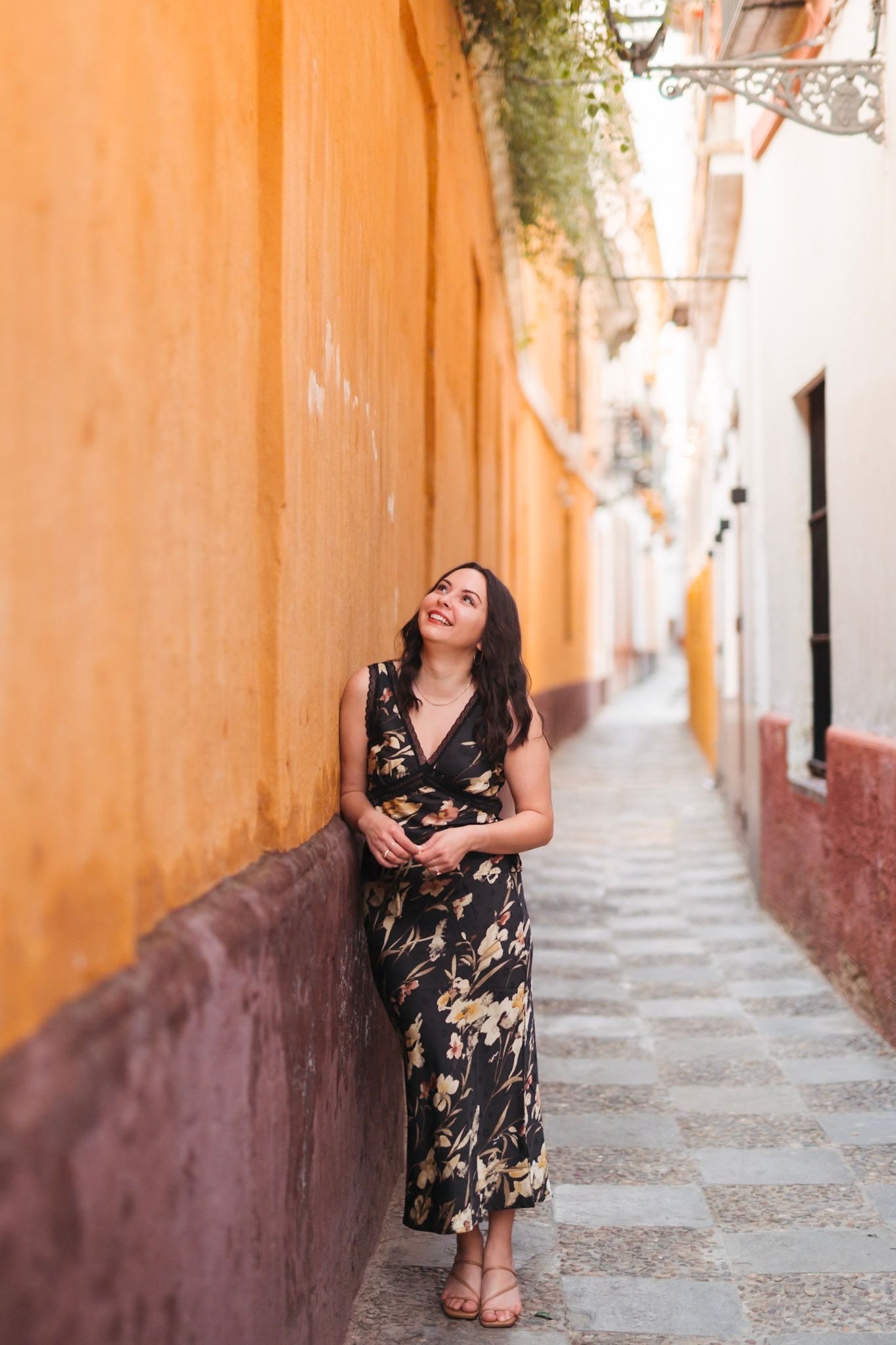 Woman in floral dress leans against orange wall in narrow alley.