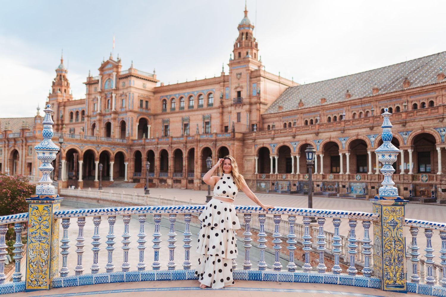 Woman in polka dot dress poses at Plaza de España, Seville, Spain, with ornate tiled balustrades and arched architecture.