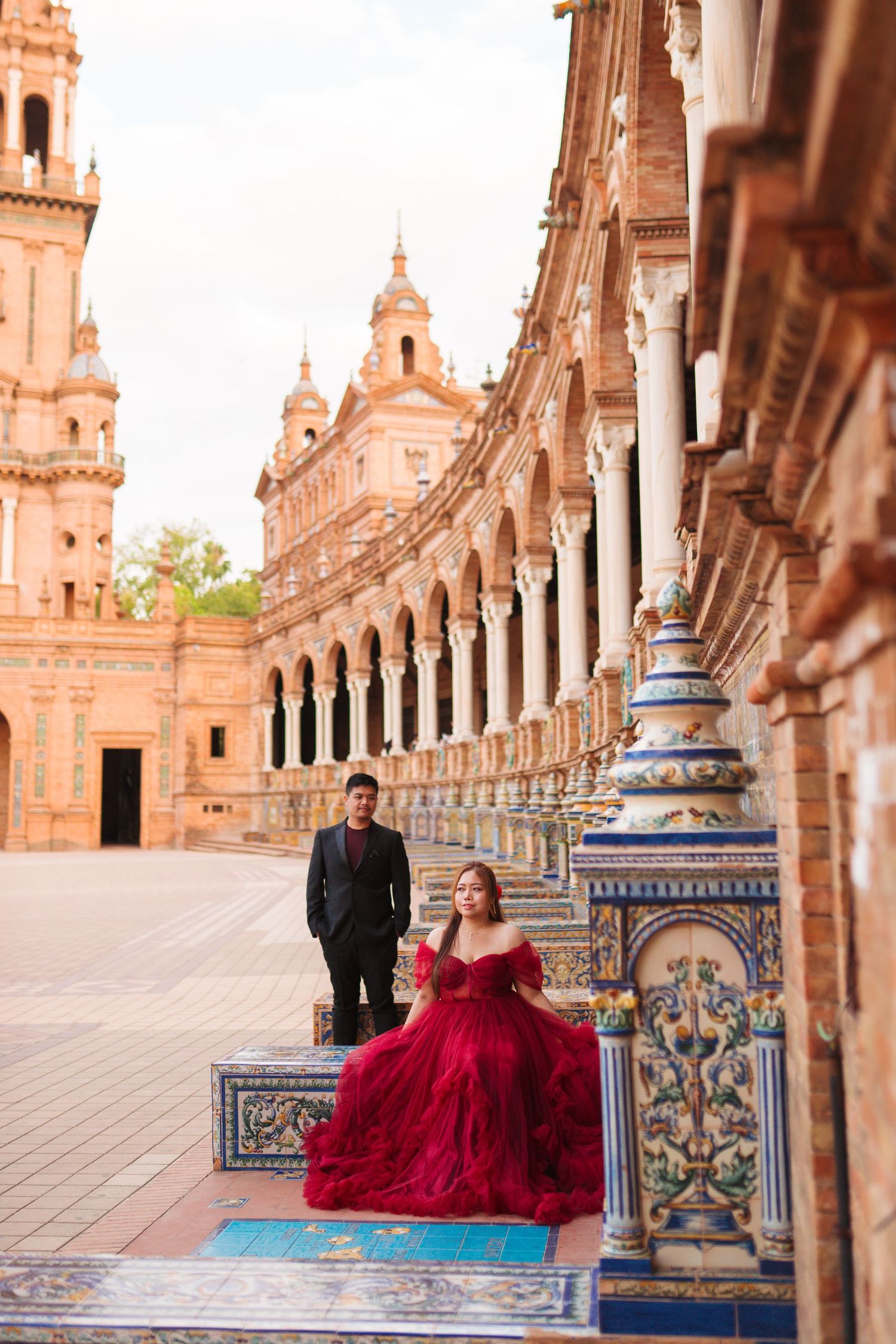 Woman in red gown poses at Plaza de España, Seville, with a man in black suit standing in the ornate arcade behind her.