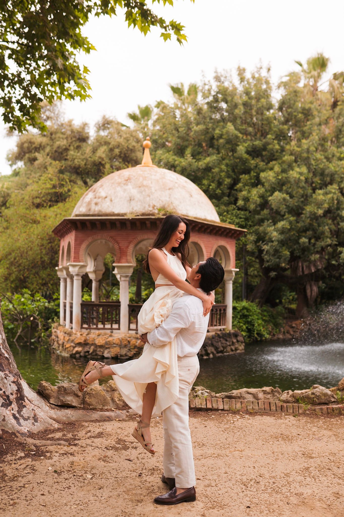 Man lifting woman joyfully near Moorish pavilion by pond in Parque de María Luisa, Seville.