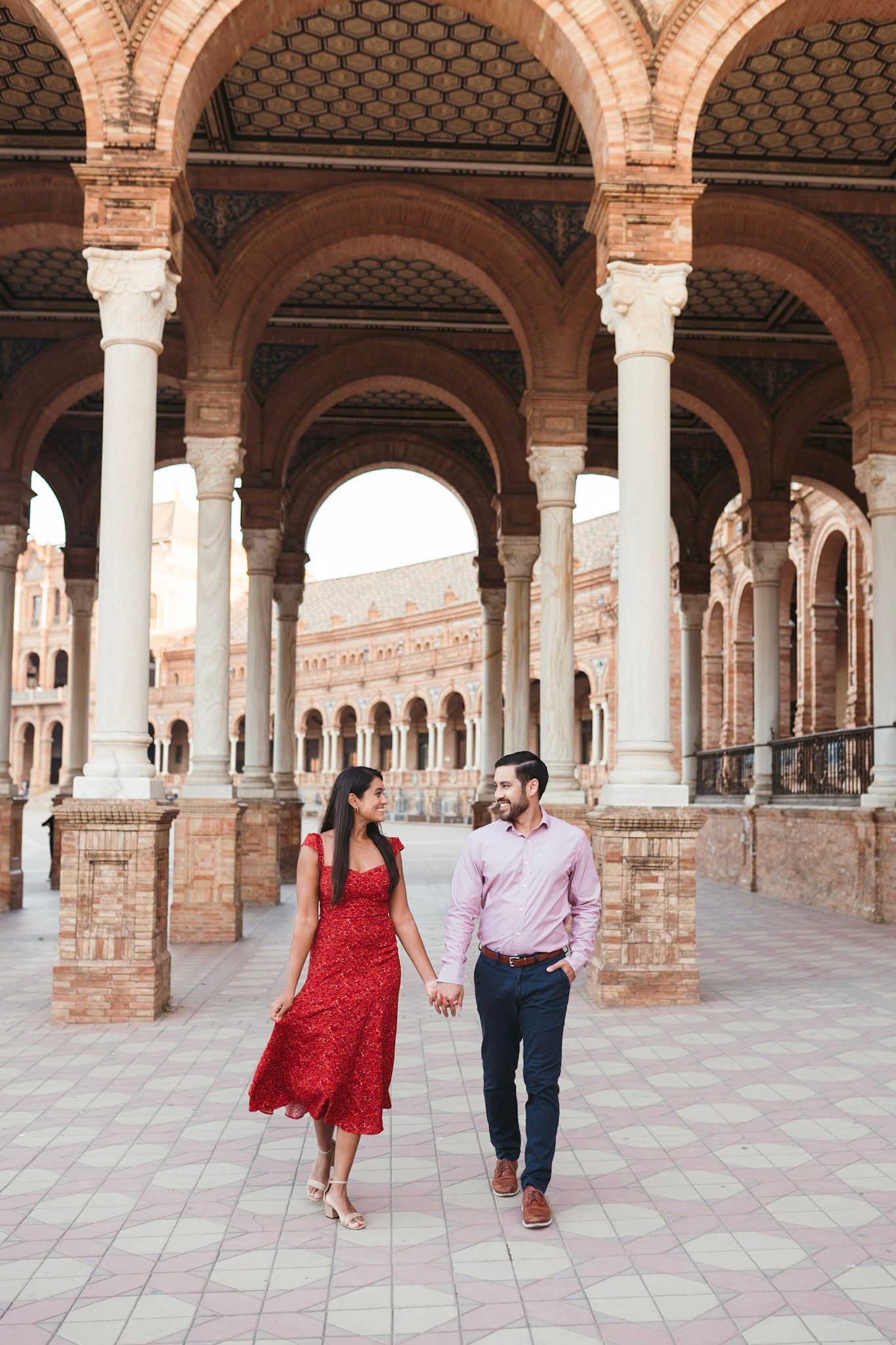 Couple holding hands and walking under arched colonnade at Plaza de España, Seville, woman in red dress.