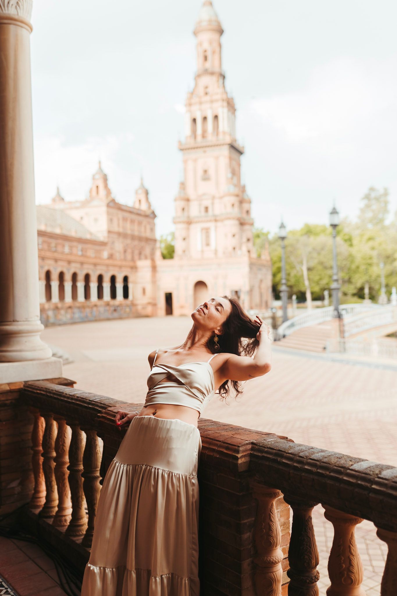 Woman in silver crop top and beige skirt posing at Plaza de España balcony with tower in background.