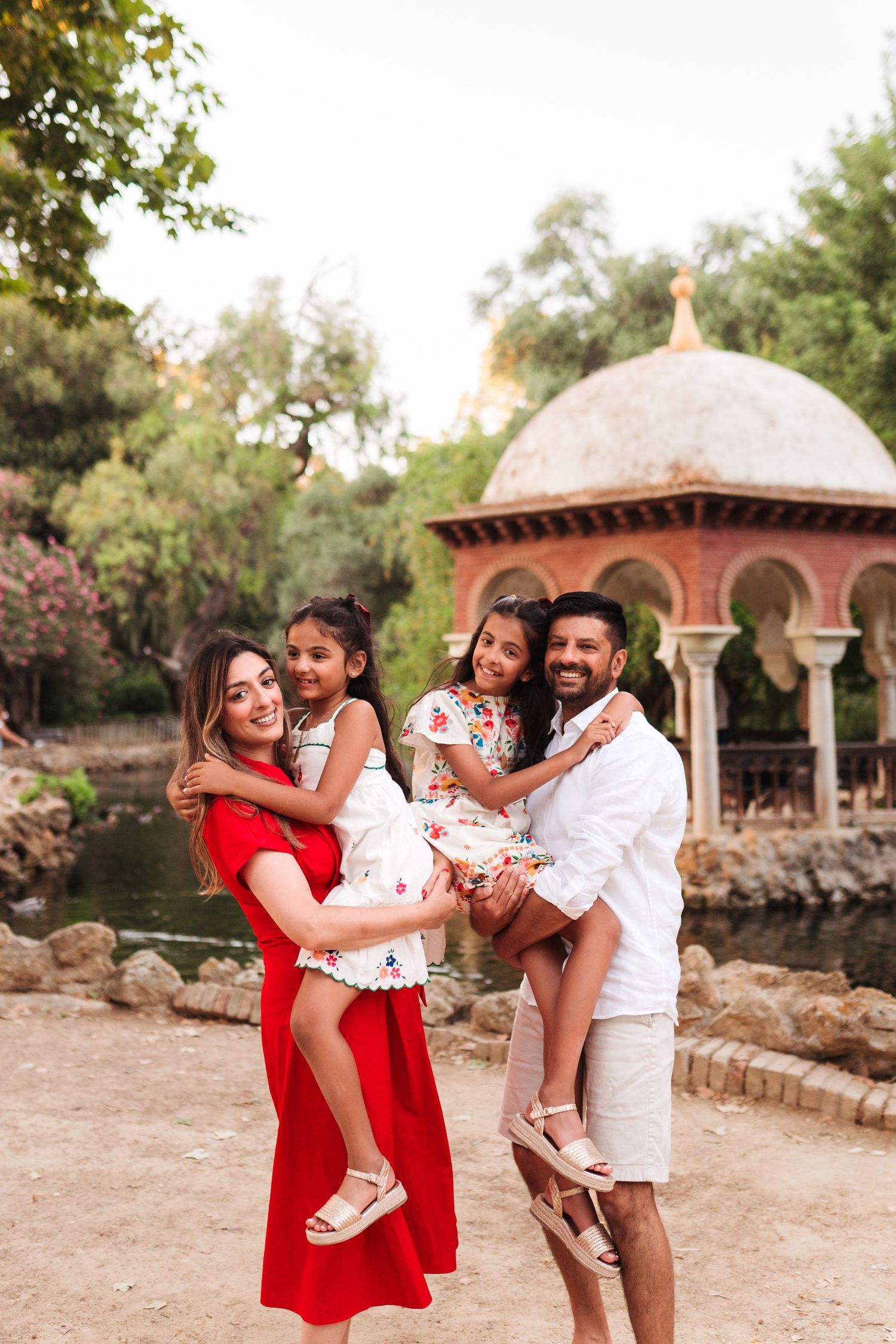 Happy family of four posing outdoors near ornate pavilion, parents holding two young daughters in white dresses at park.
