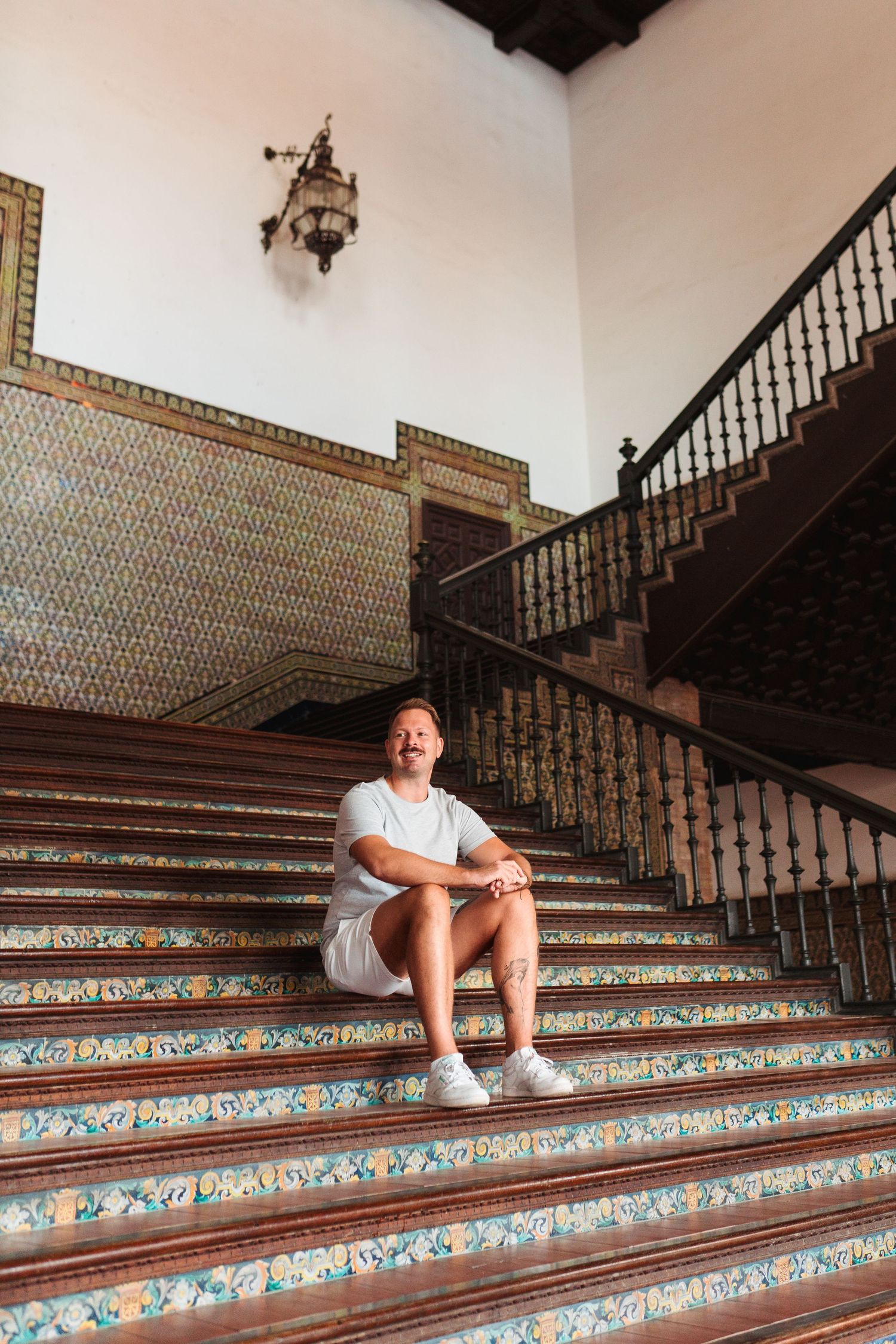 Man in white outfit sitting on ornate tiled staircase with wrought iron railing in historic building.
