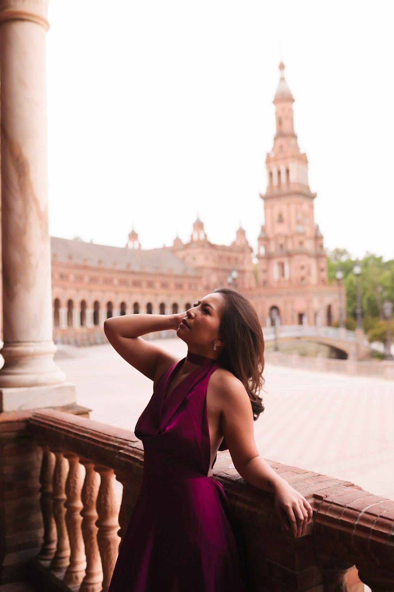Elegant woman in purple gown leans on balustrade at Plaza de España, Seville, gazing upward dreamily.