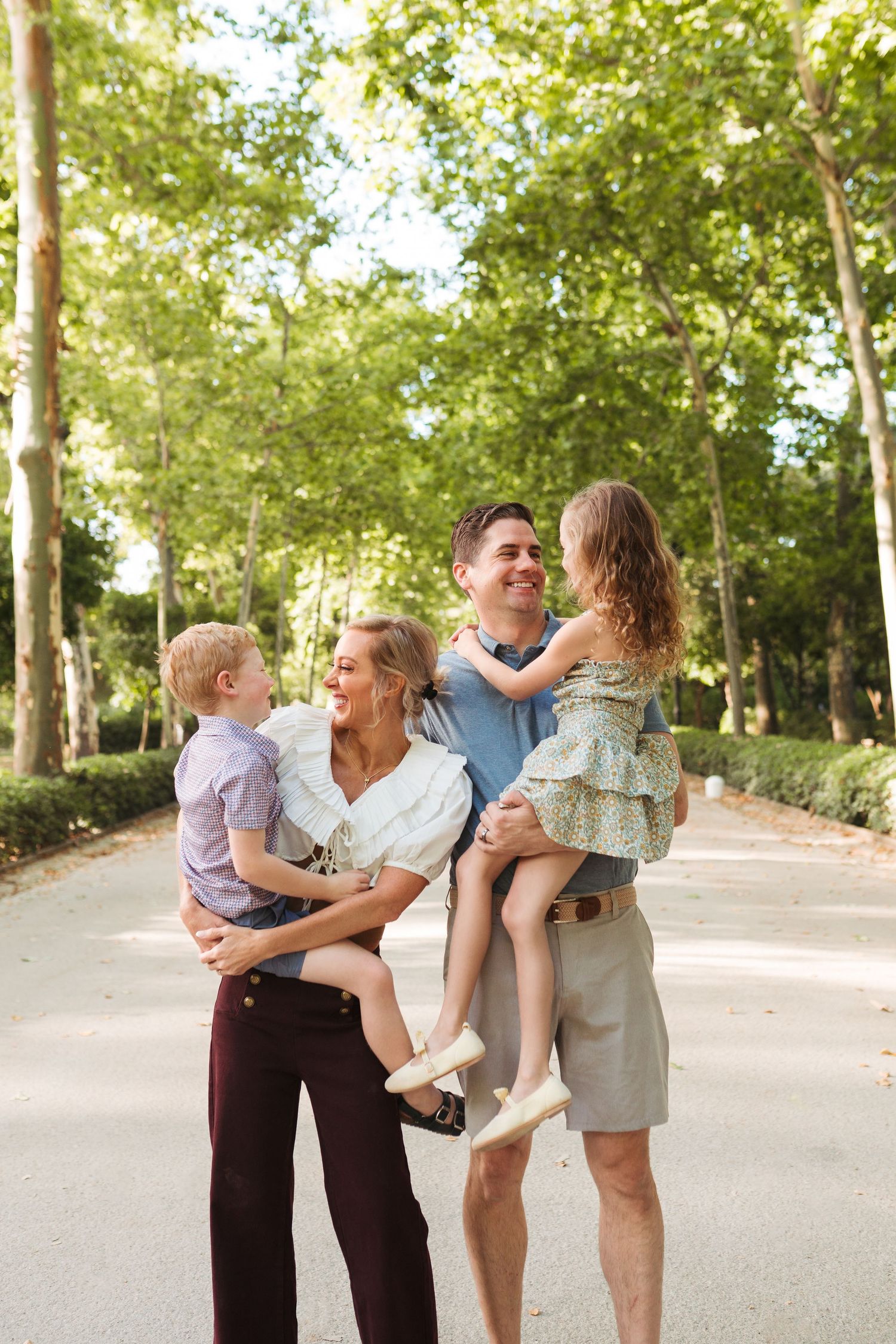 Happy family of four enjoying a sunny day in a tree-lined park, parents holding their young children while laughing together.