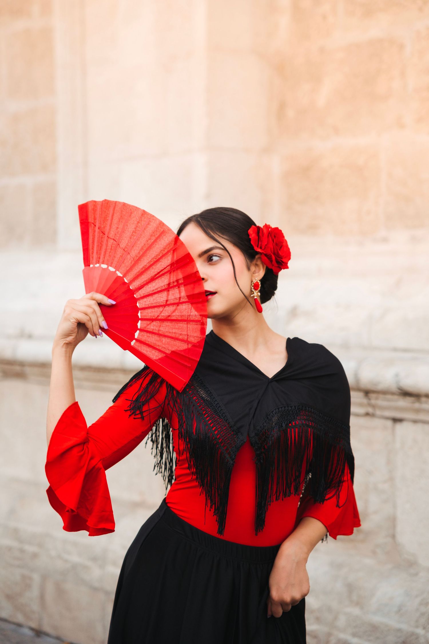 Flamenco dancer in red and black dress holding red fan against stone wall.