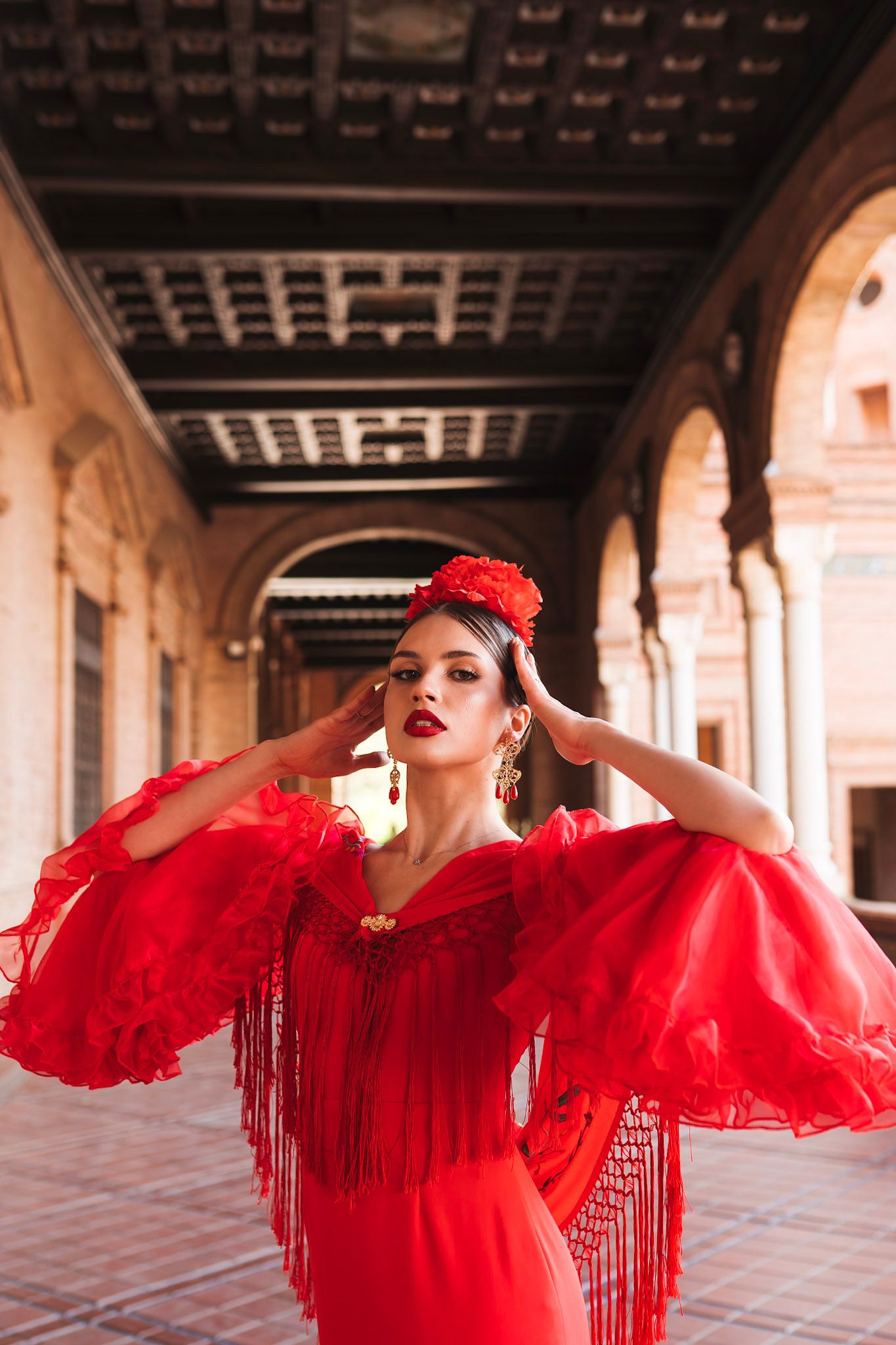 Elegant flamenco dancer in red fringed dress with flower hair accessory posing in ornate Spanish arcade.