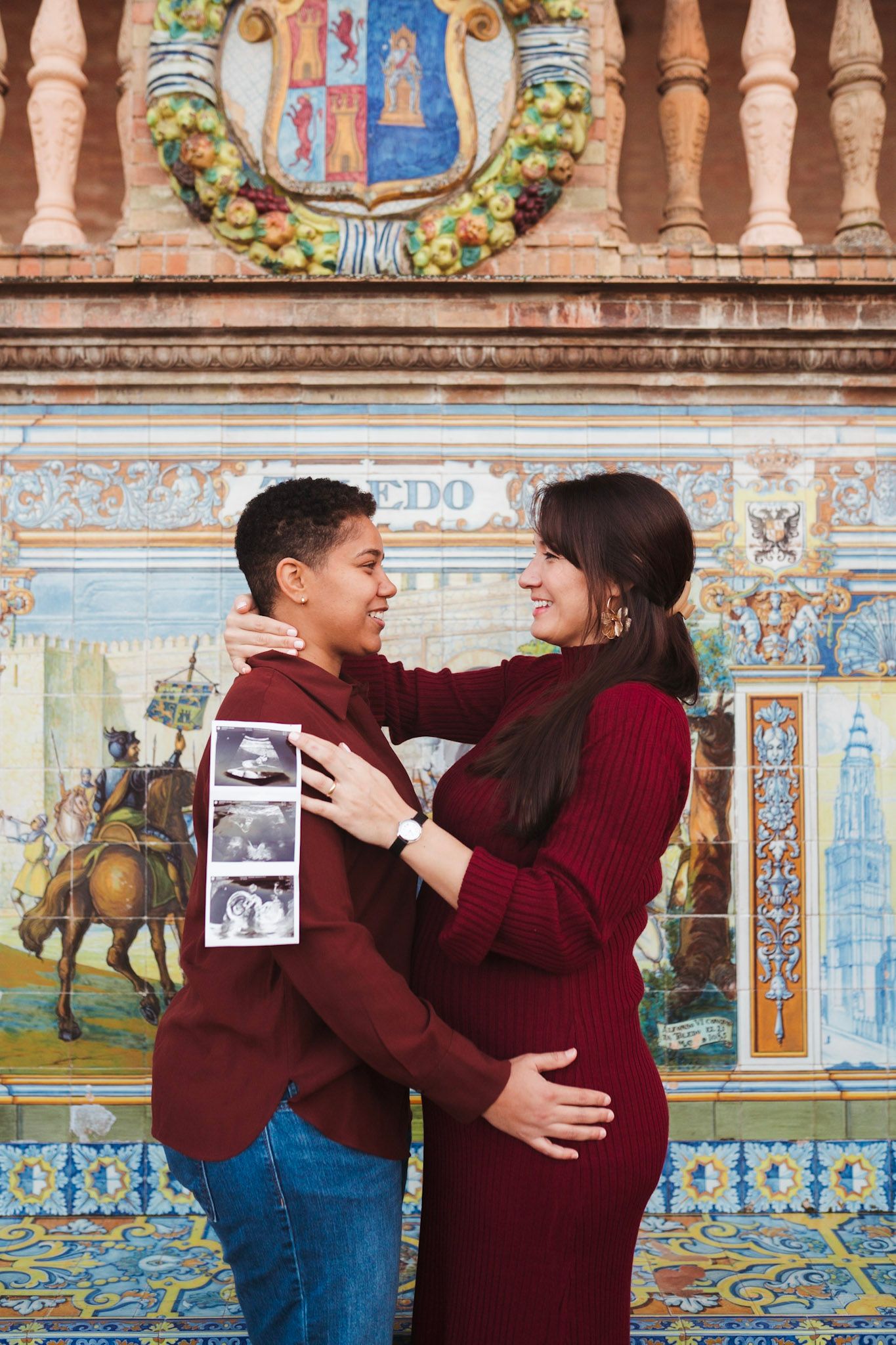 Happy expecting couple embraces holding ultrasound images before ornate Toledo tile panel in Spain.