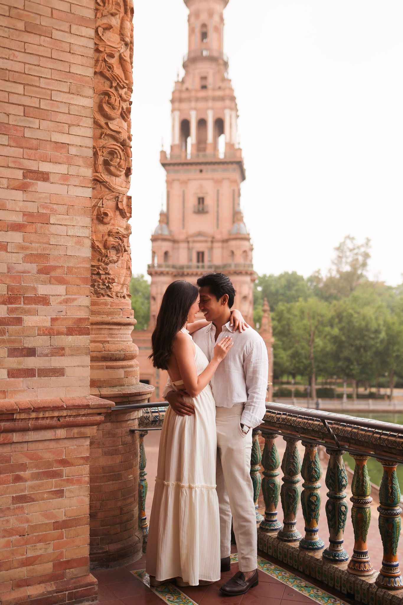 Romantic couple embracing on ornate balcony at Plaza de España, Seville, with iconic brick tower in background.