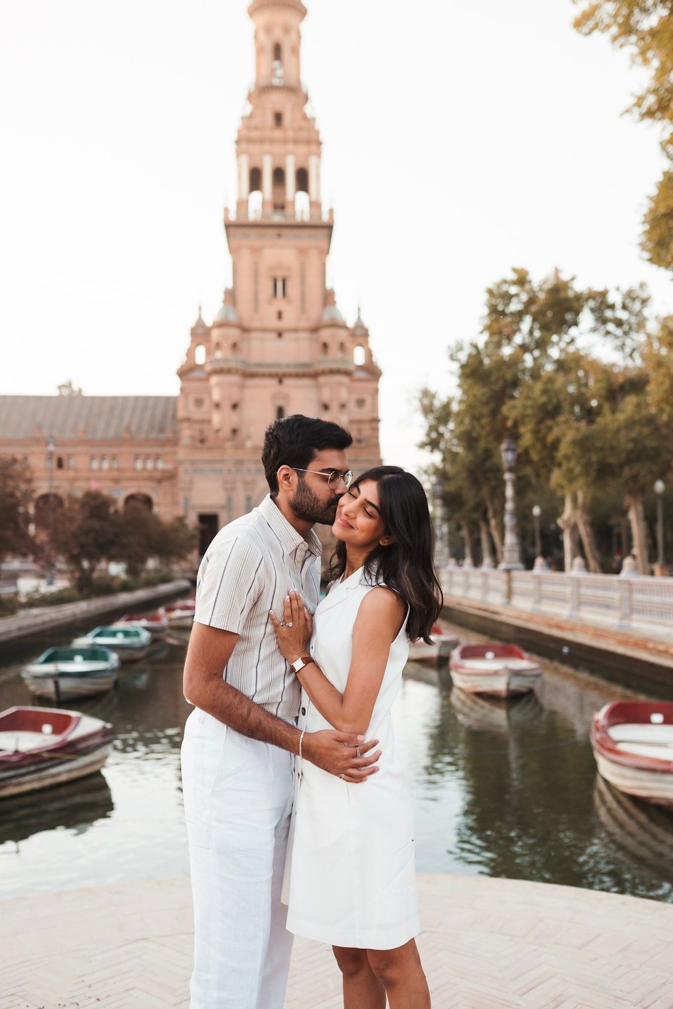 South Asian couple embracing near canal at Plaza de España, Seville, with boats and tower in background.