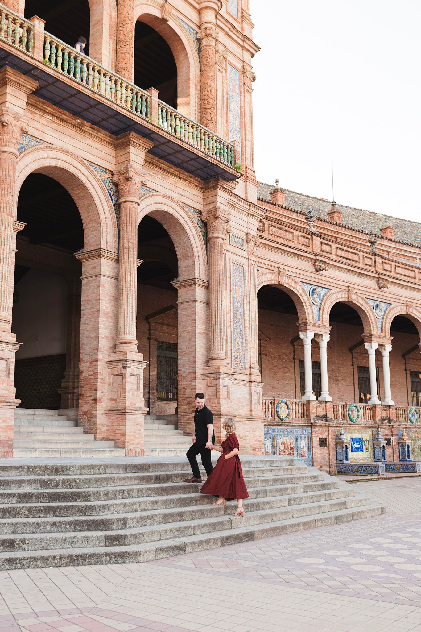 Couple walking down grand stone steps of Plaza de Espana in Seville, surrounded by stunning brick archways.