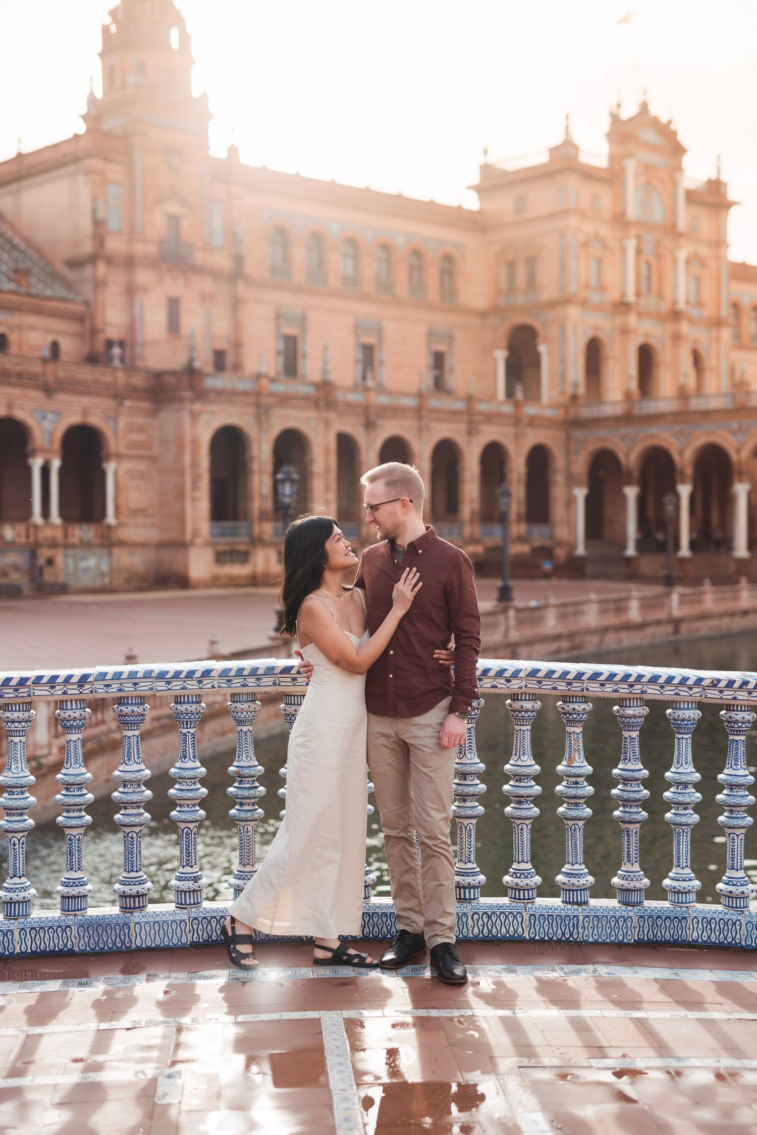 Couple embracing on a rain-soaked tiled bridge at Plaza de España, Seville, bathed in warm sunset light.