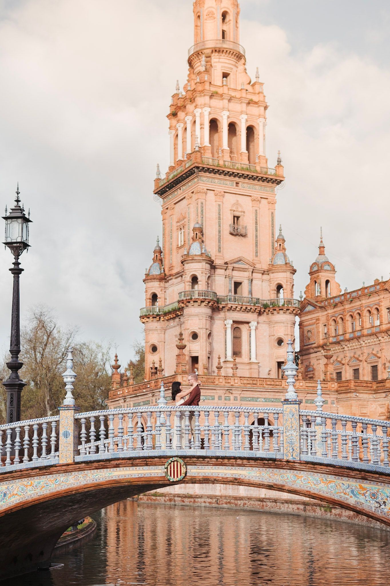 Couple sitting on a tiled bridge at Plaza de España, Seville, with a towering baroque spire reflected below.