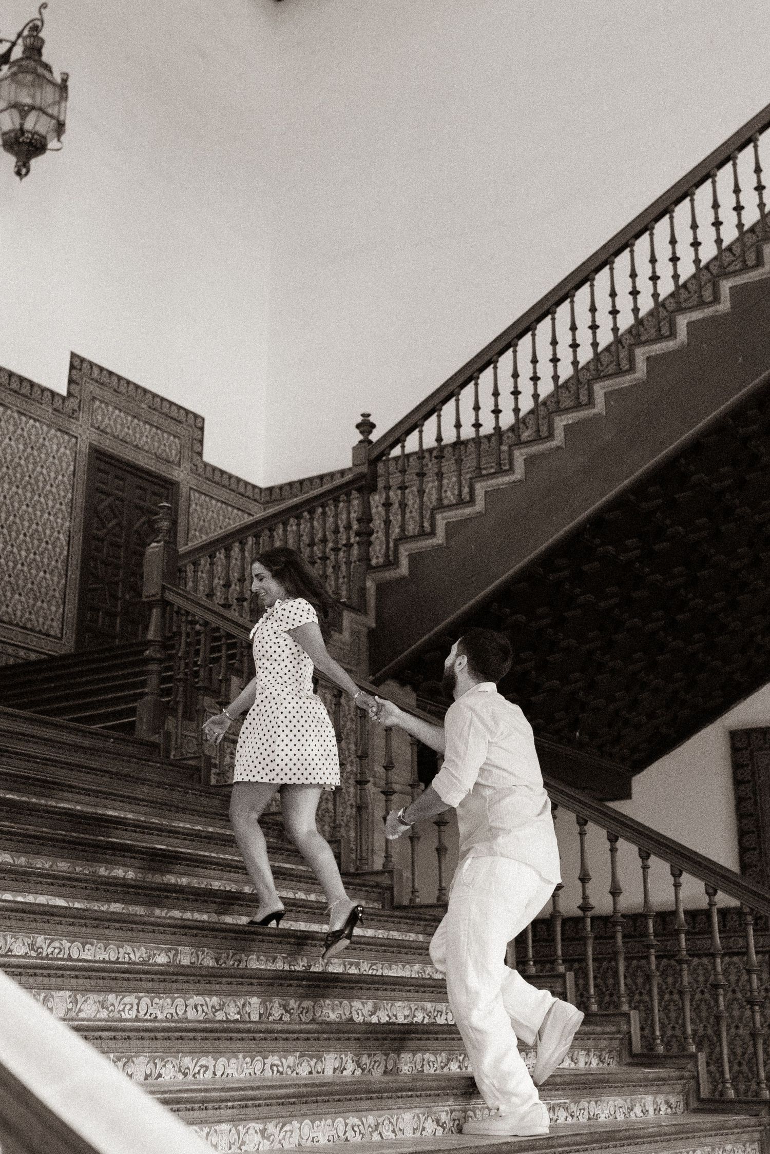 Black and white photo of a couple holding hands while climbing an ornate staircase with decorative railings.