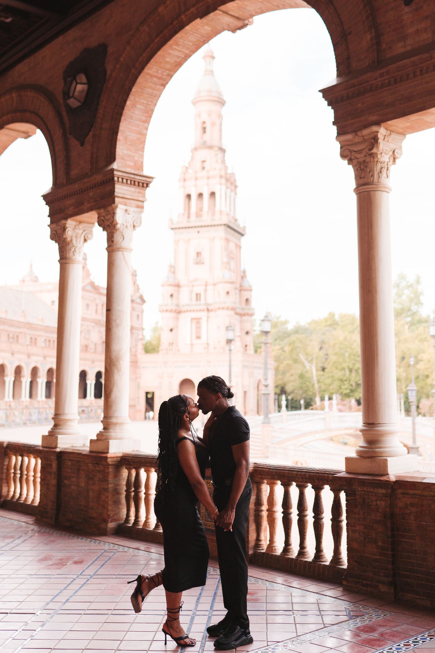 Couple sharing a romantic kiss under ornate arches at Plaza de España, Seville, with a grand tower in the background.