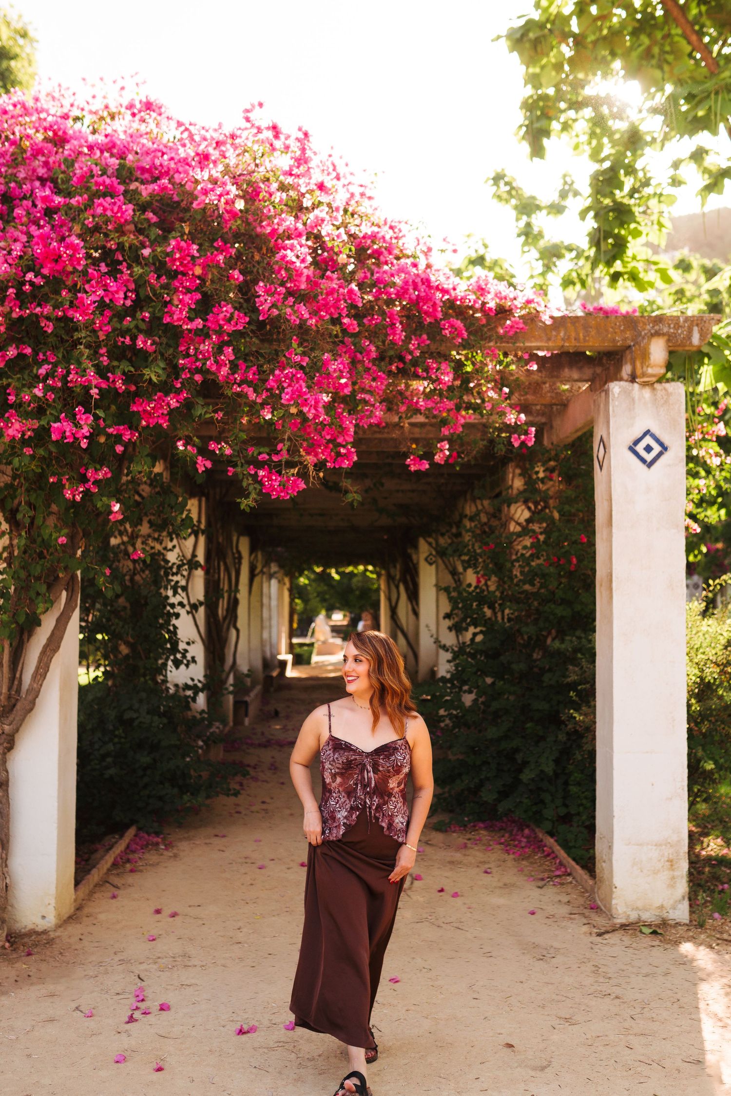 Woman in burgundy dress walks under bougainvillea-covered pergola with vibrant pink flowers.