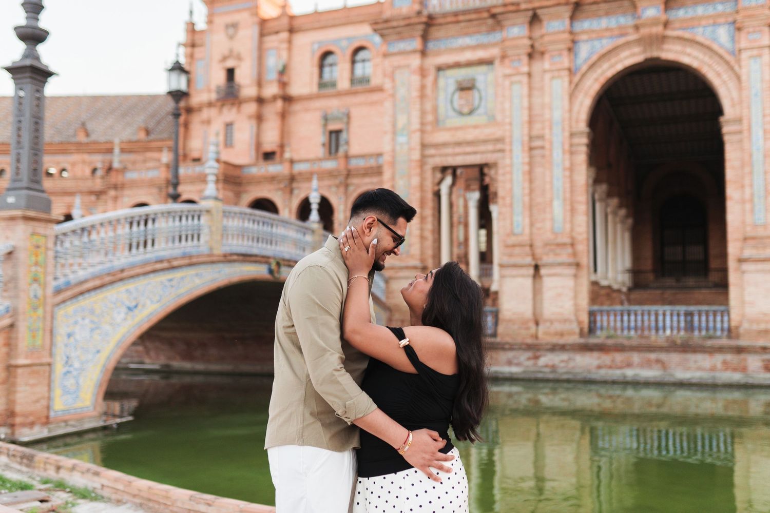Romantic couple sharing a kiss at Plaza de España in Seville, Spain, with a scenic bridge and green canal in the background.