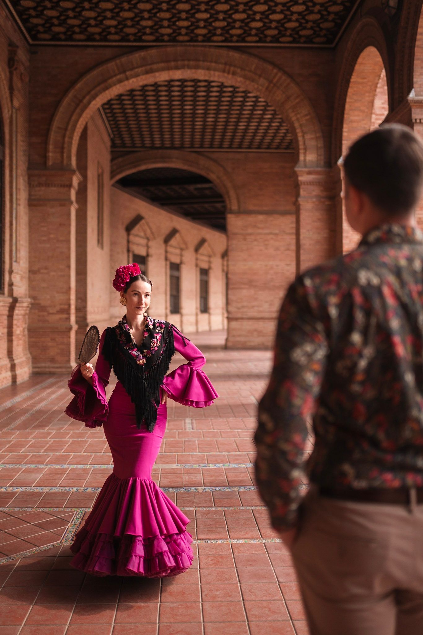 Flamenco dancer in pink ruffled dress holding fan poses in brick archway corridor while man watches.