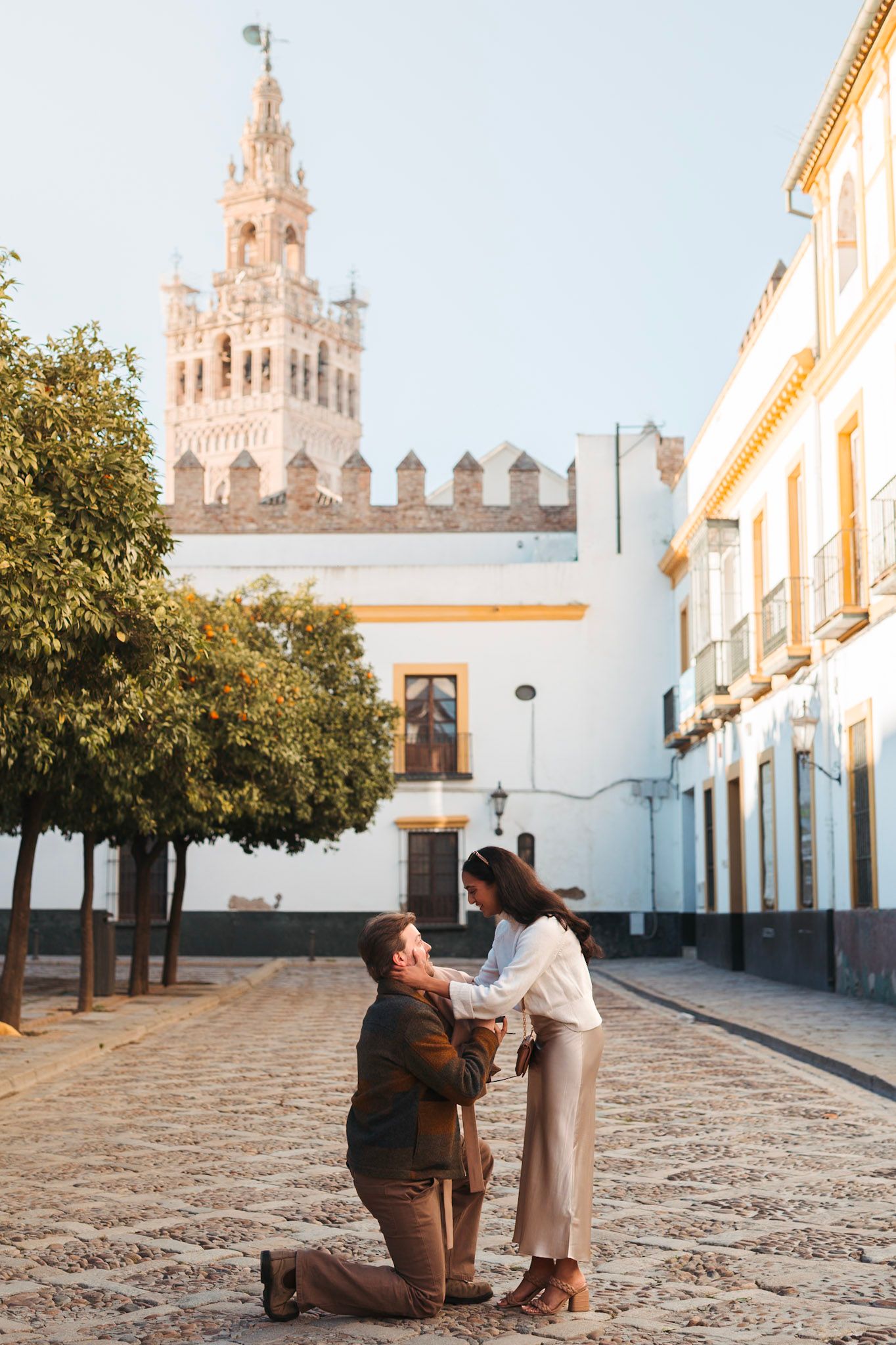 Man on bended knee proposing marriage to woman near Seville cathedral Giralda tower at sunrise.