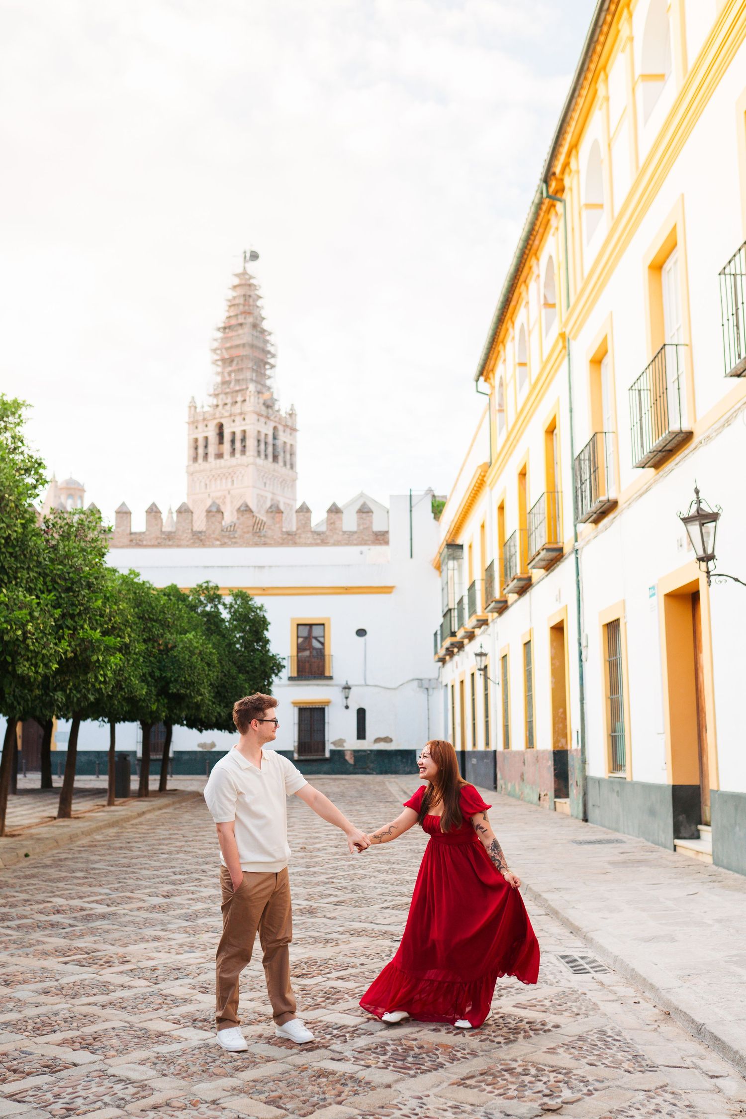Couple holding hands in red dress and casual wear on cobblestone street with historic tower in Seville, Spain.