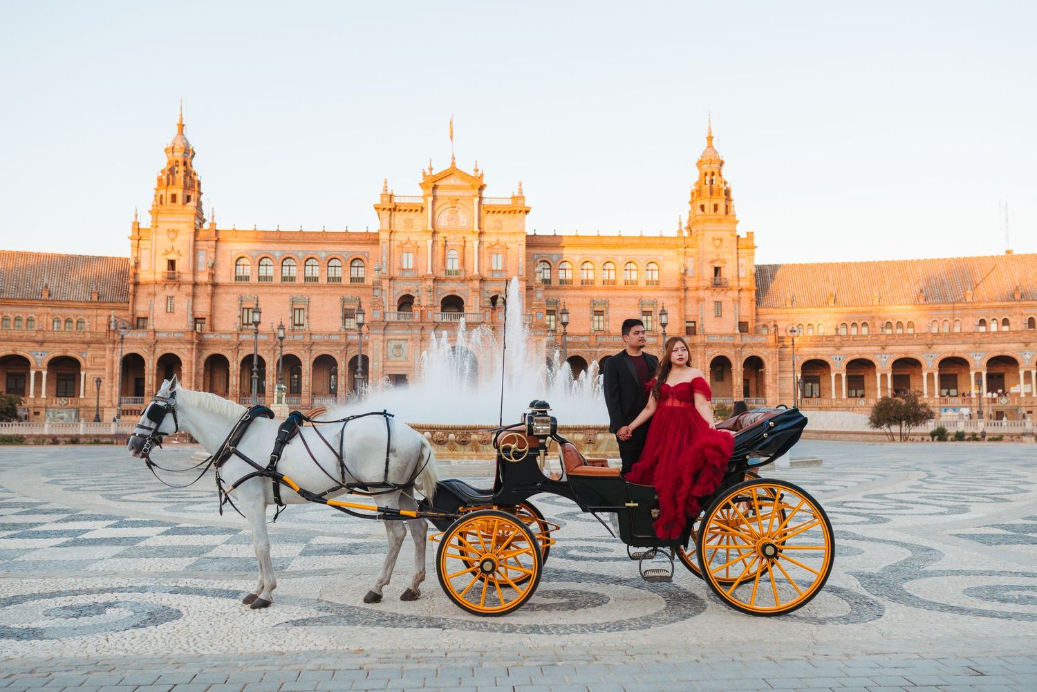 Couple in formal attire riding a golden horse-drawn carriage at Plaza de España, Seville, Spain at sunset.