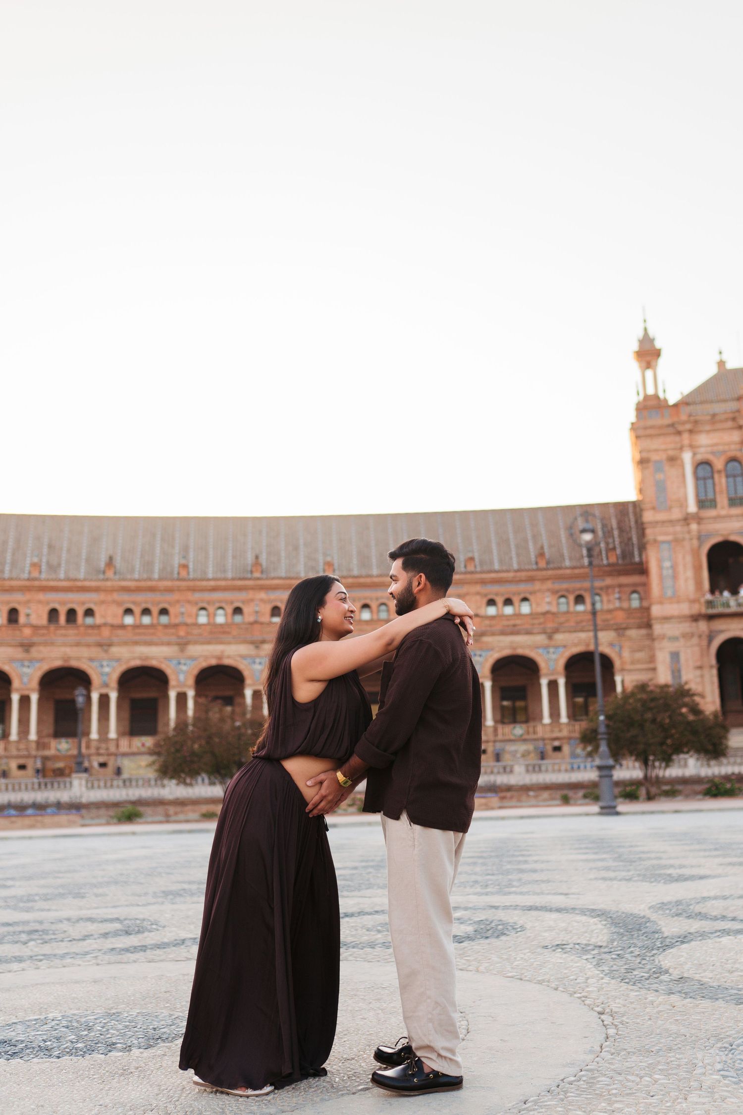 Couple embracing romantically at Plaza de España in Seville, Spain, dressed in elegant dark attire at sunset.