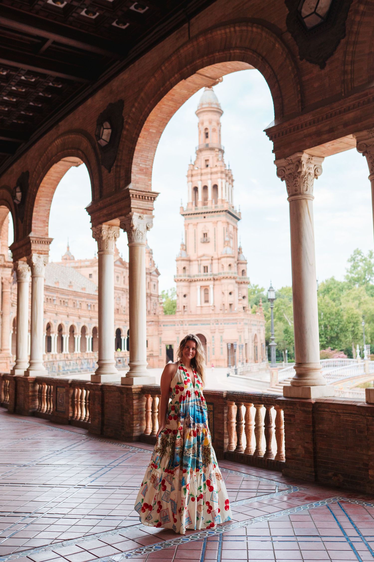 Woman in floral maxi dress posing under arched colonnade at Plaza de España, Seville, Spain.