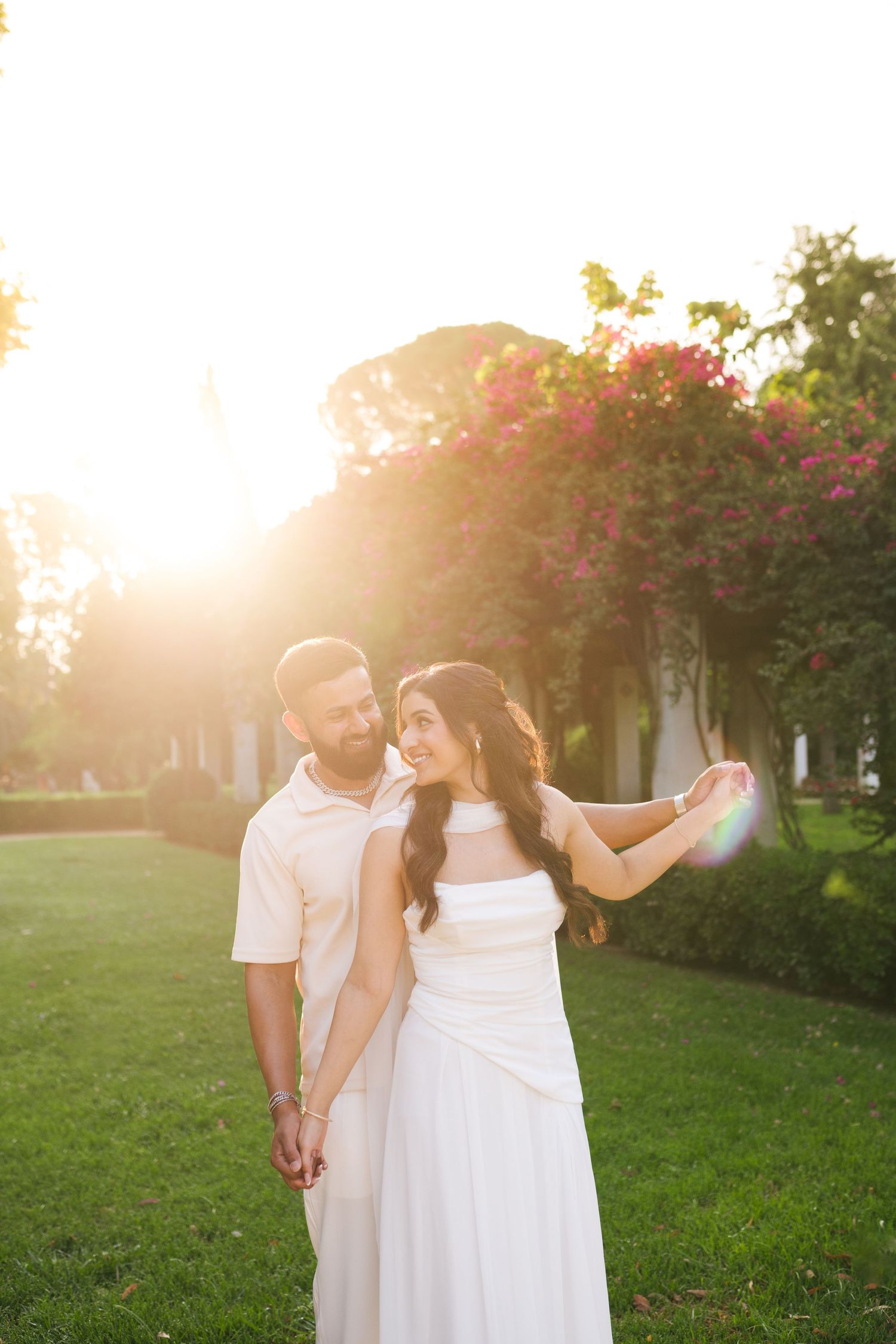 Couple holding hands in golden sunset light, woman in white dress with pink flowers and green lawn backdrop.
