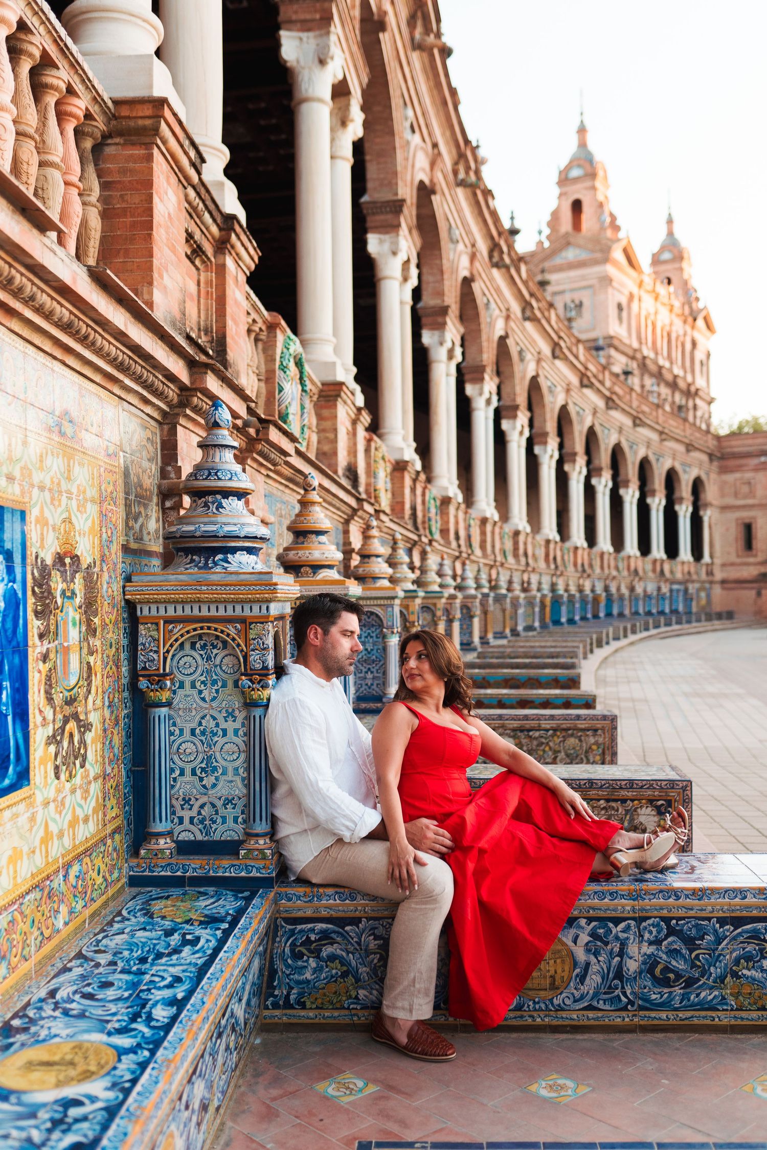 Couple sitting on ornate tiled bench at Plaza de España, Seville, woman in red dress, man in white shirt.