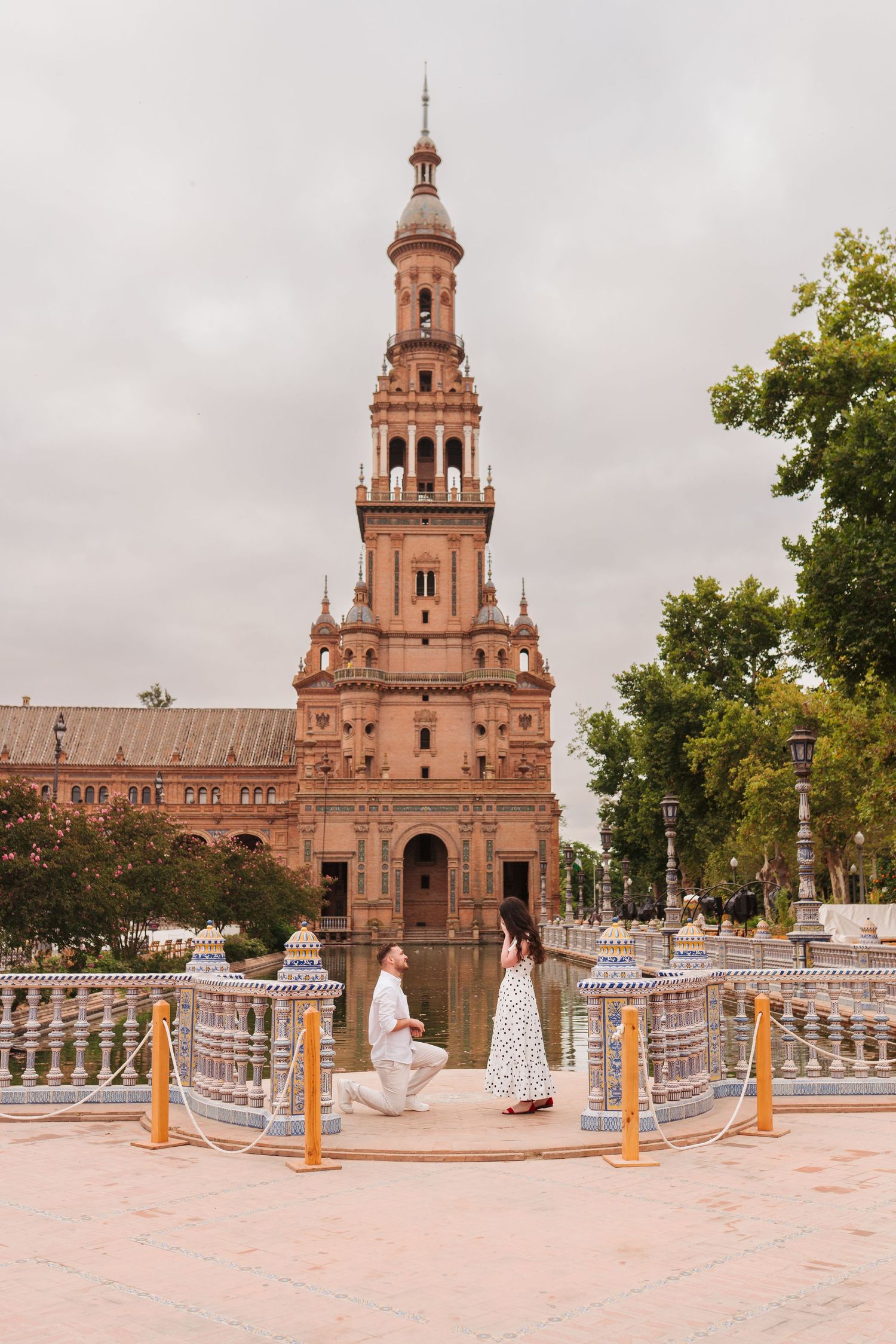 Man proposing to woman at Plaza de España in Seville, Spain, with iconic baroque tower and canal in background.