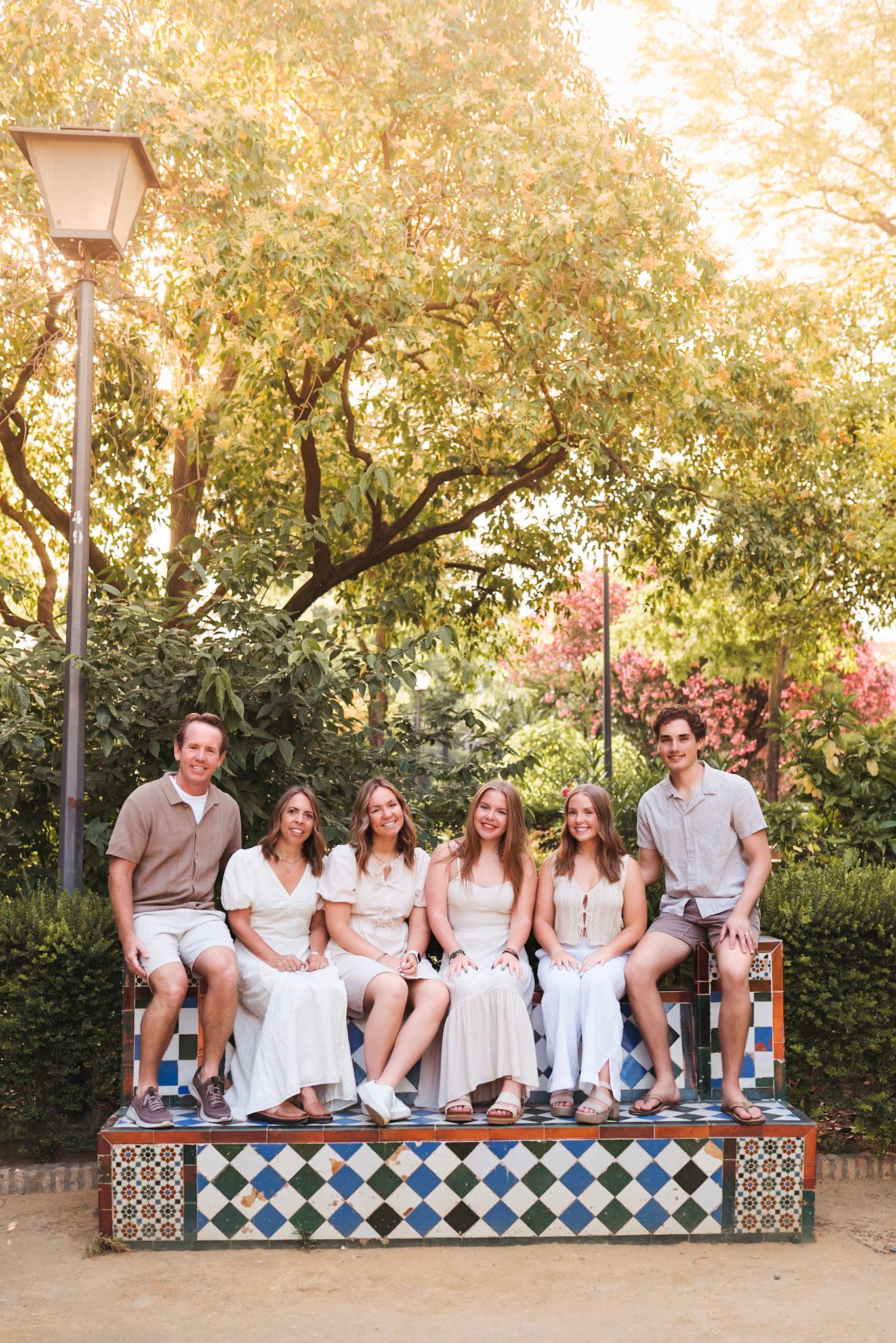 Family of six smiling on decorative tiled bench surrounded by lush trees in Seville.