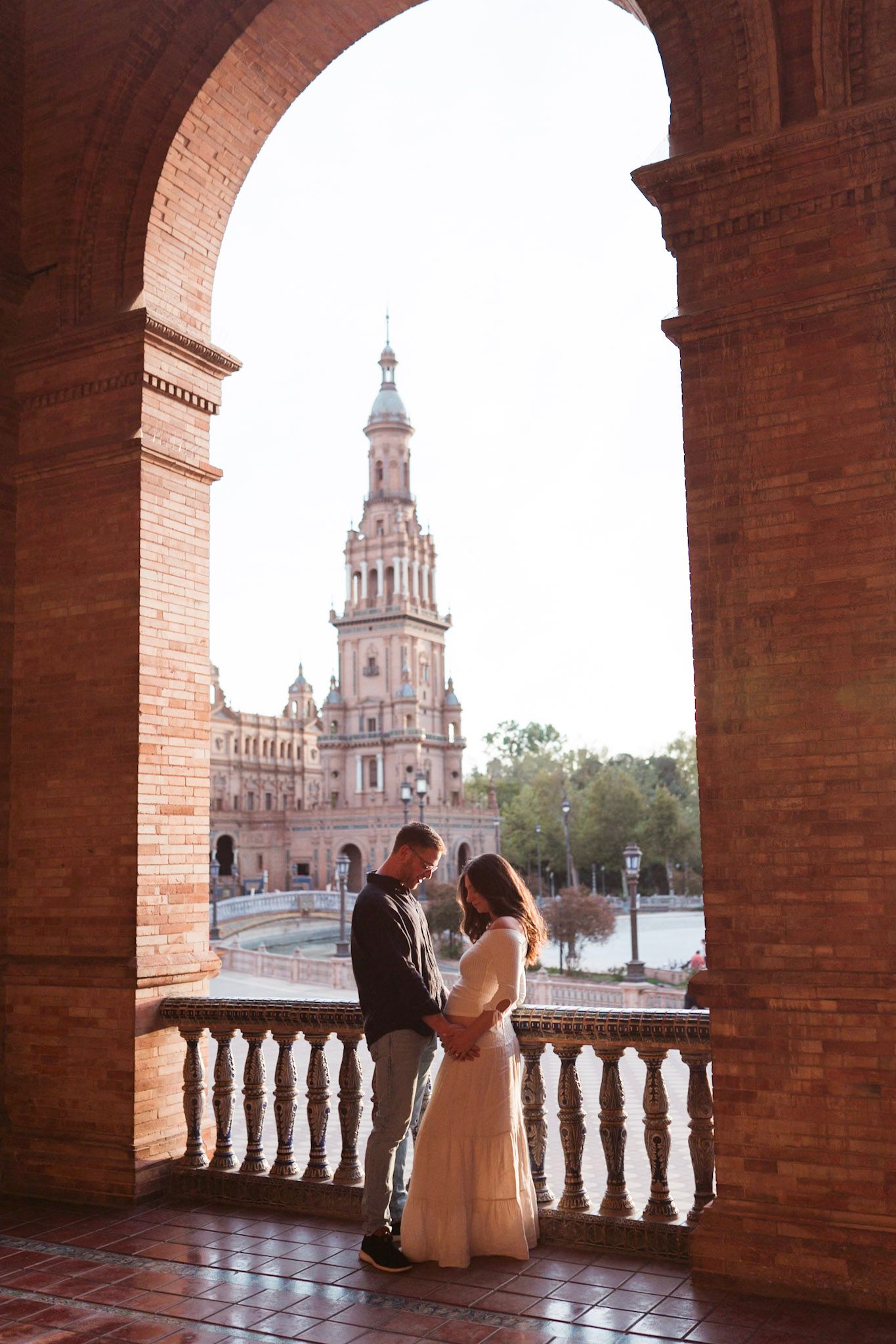 Pregnant couple embraces under brick arch with Plaza de España tower backdrop.