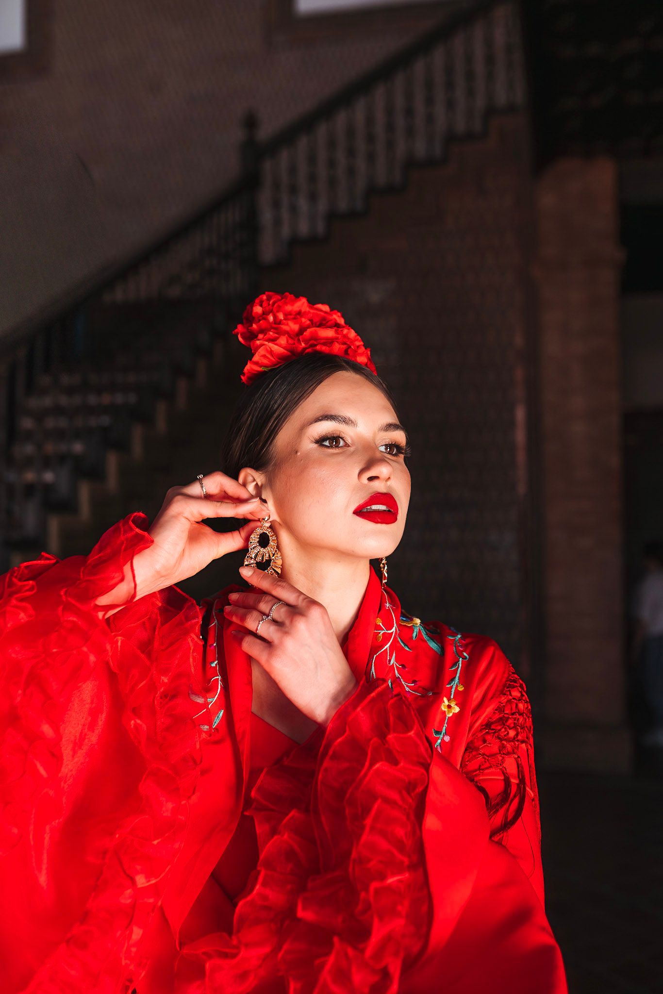Beautiful flamenco dancer in red dress adjusting gold earring, red flower hair accessory, dramatic lighting.