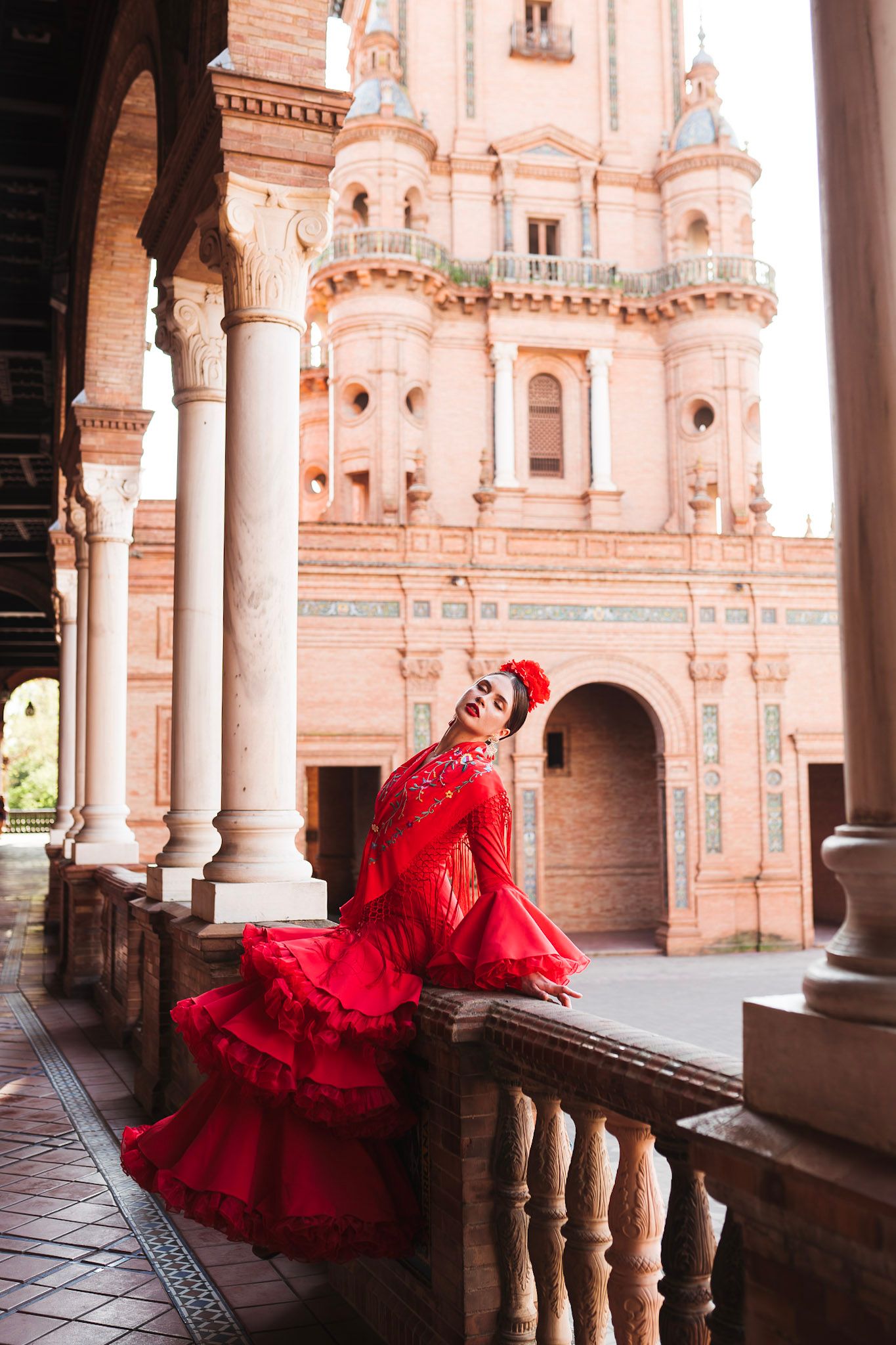 Flamenco dancer in red ruffled gown posing gracefully on balcony of Plaza de España, Seville.