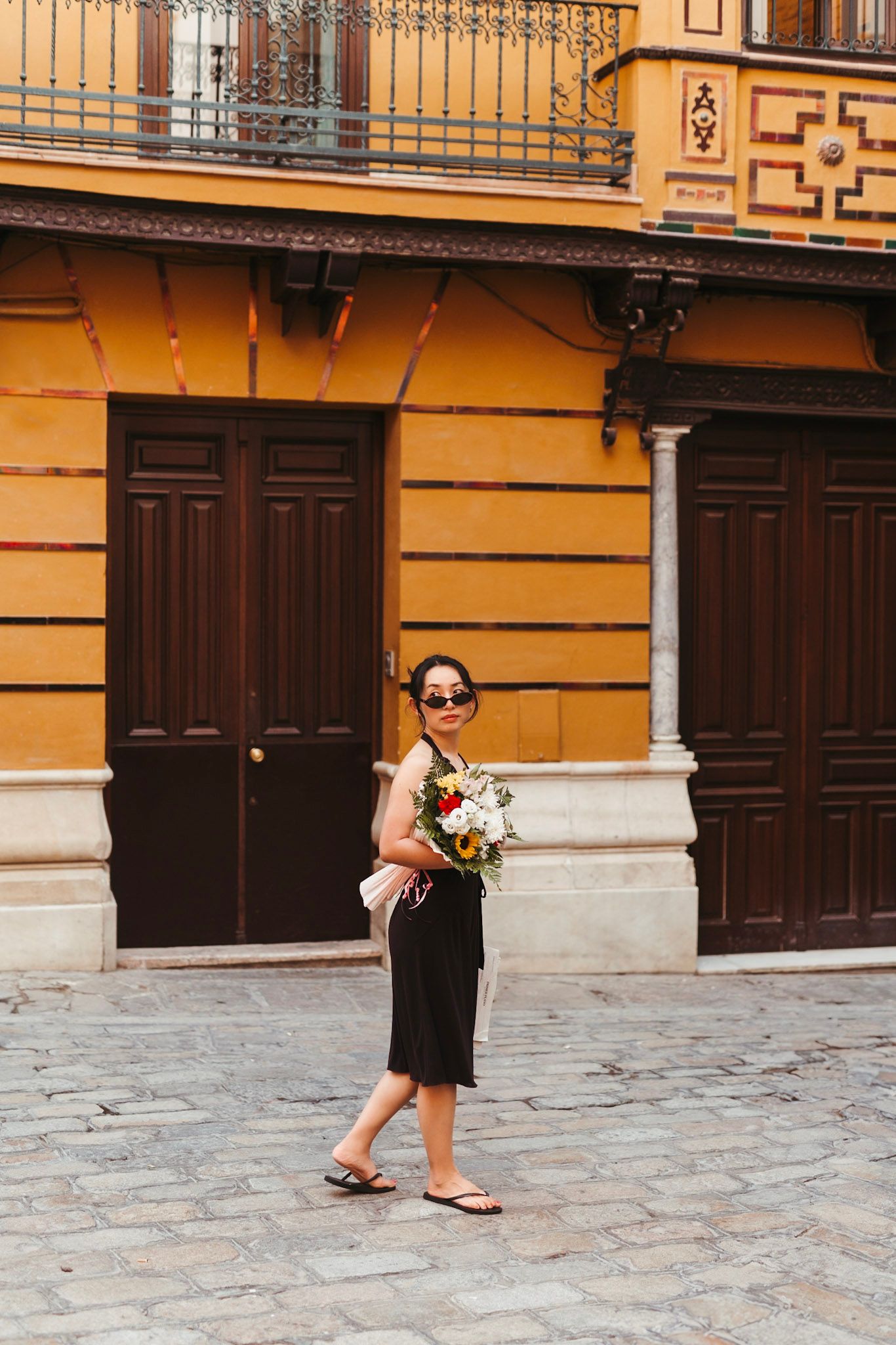 Asian woman in black skirt holding colorful flower bouquet walking past orange building in Seville.