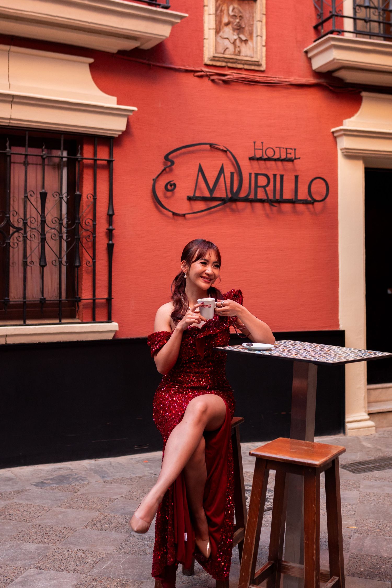 Elegant woman in red sequin dress sitting at outdoor cafe table at Hotel Murillo Seville.