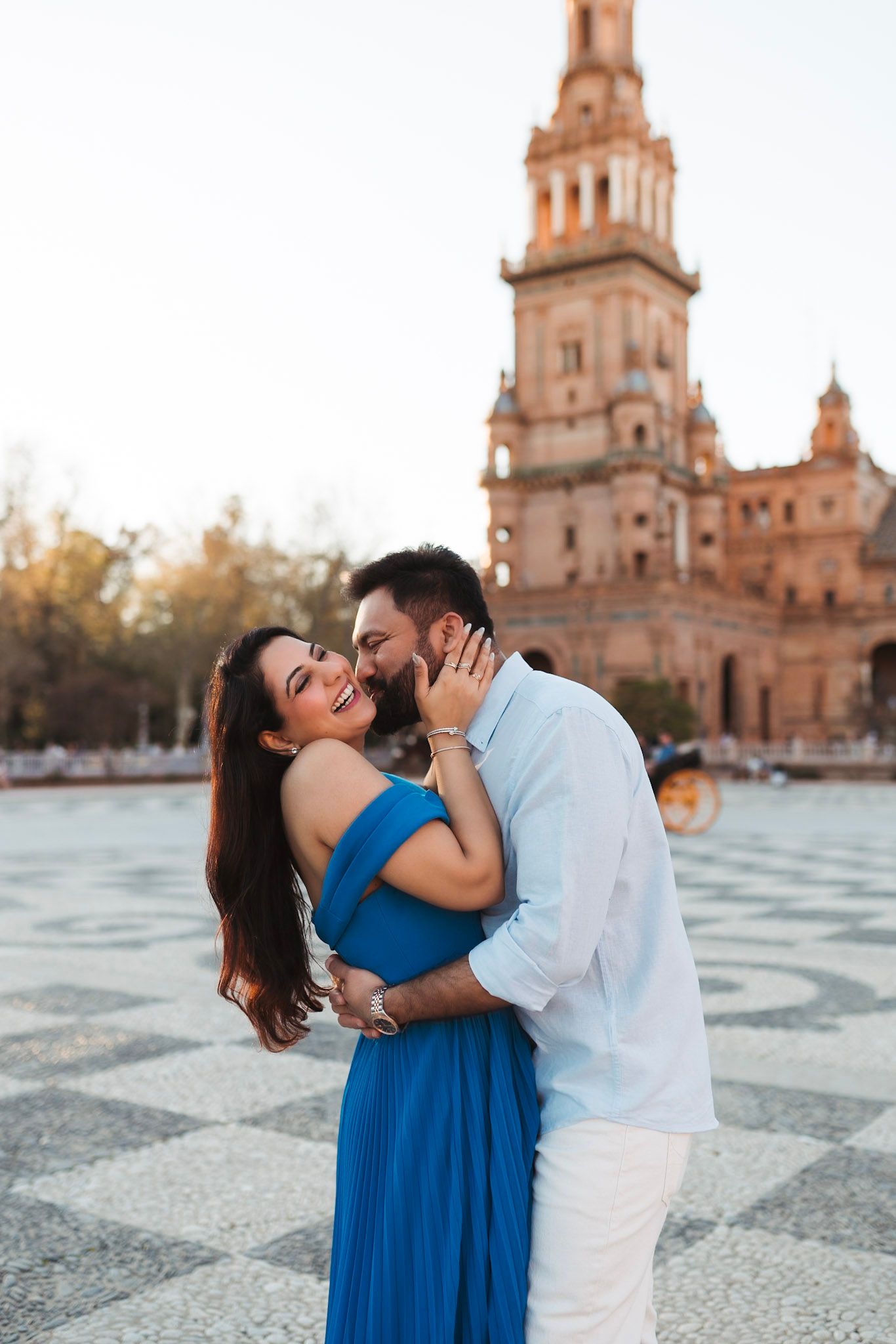 Joyful couple embracing and laughing at sunset in Plaza de España, Seville, woman in blue dress.