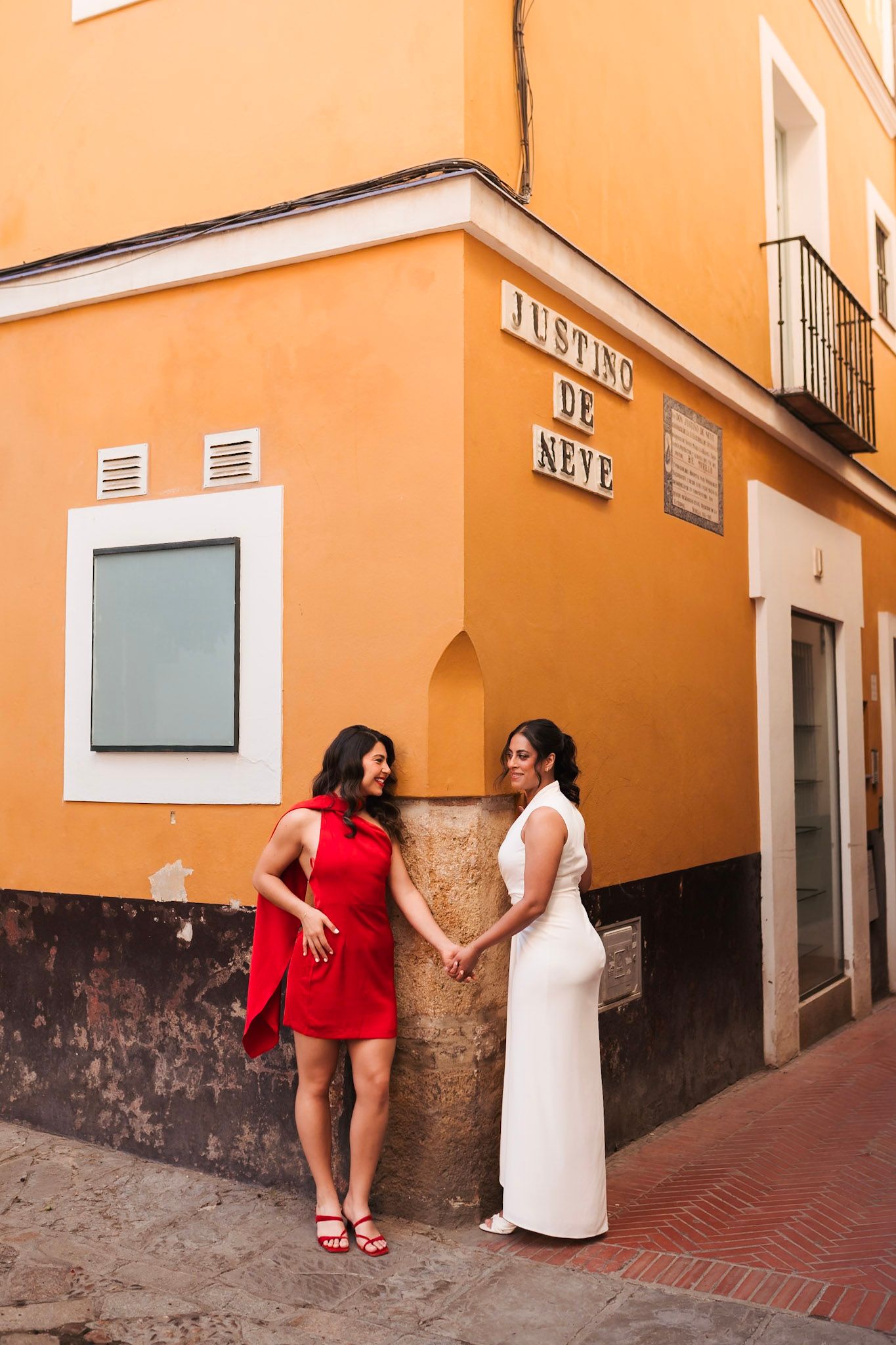 Two women in red and white dresses hold hands by a colorful orange building in Seville, Spain.