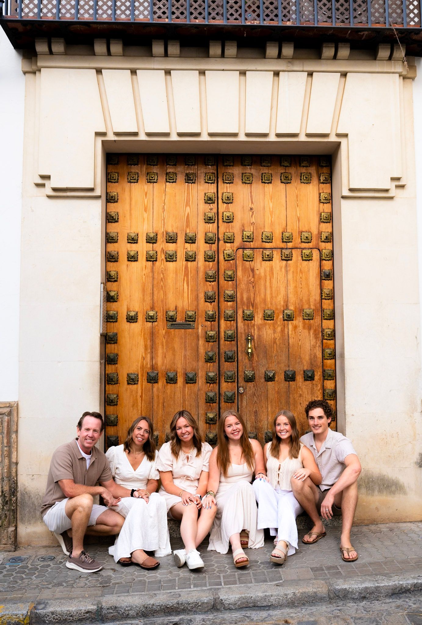 Family of six in white outfits seated before large wooden studded door in Seville.