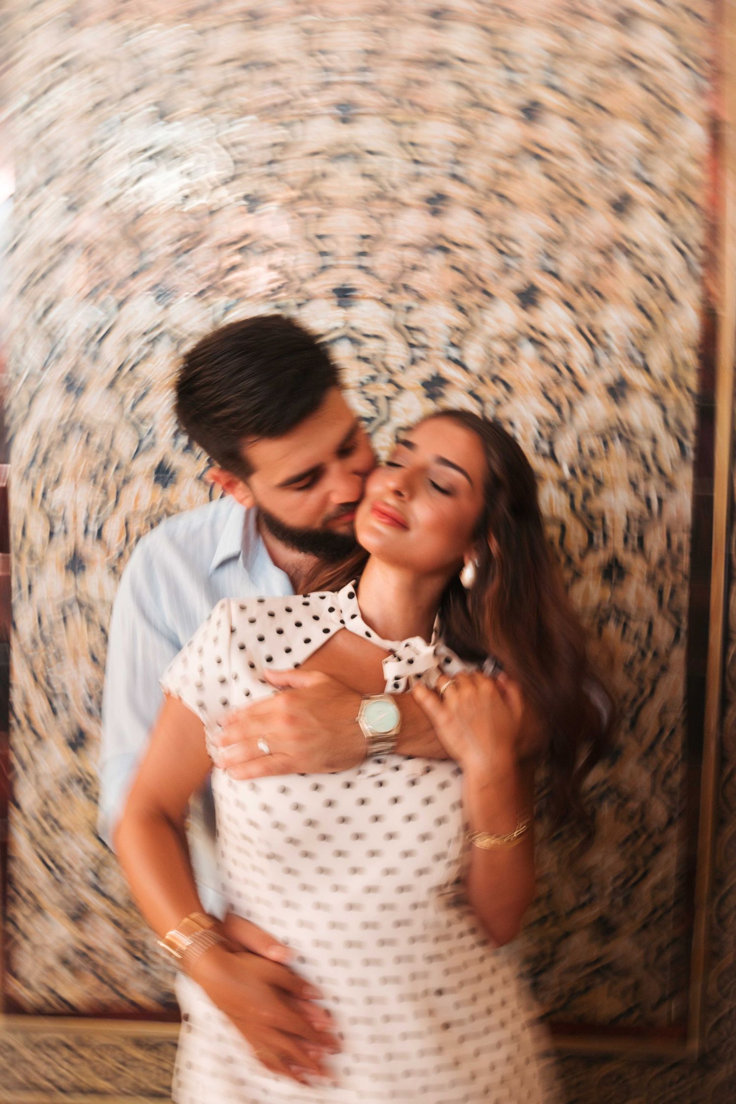 A couple embraces tenderly, the man kissing the woman's cheek as she closes her eyes, standing before a patterned wall.