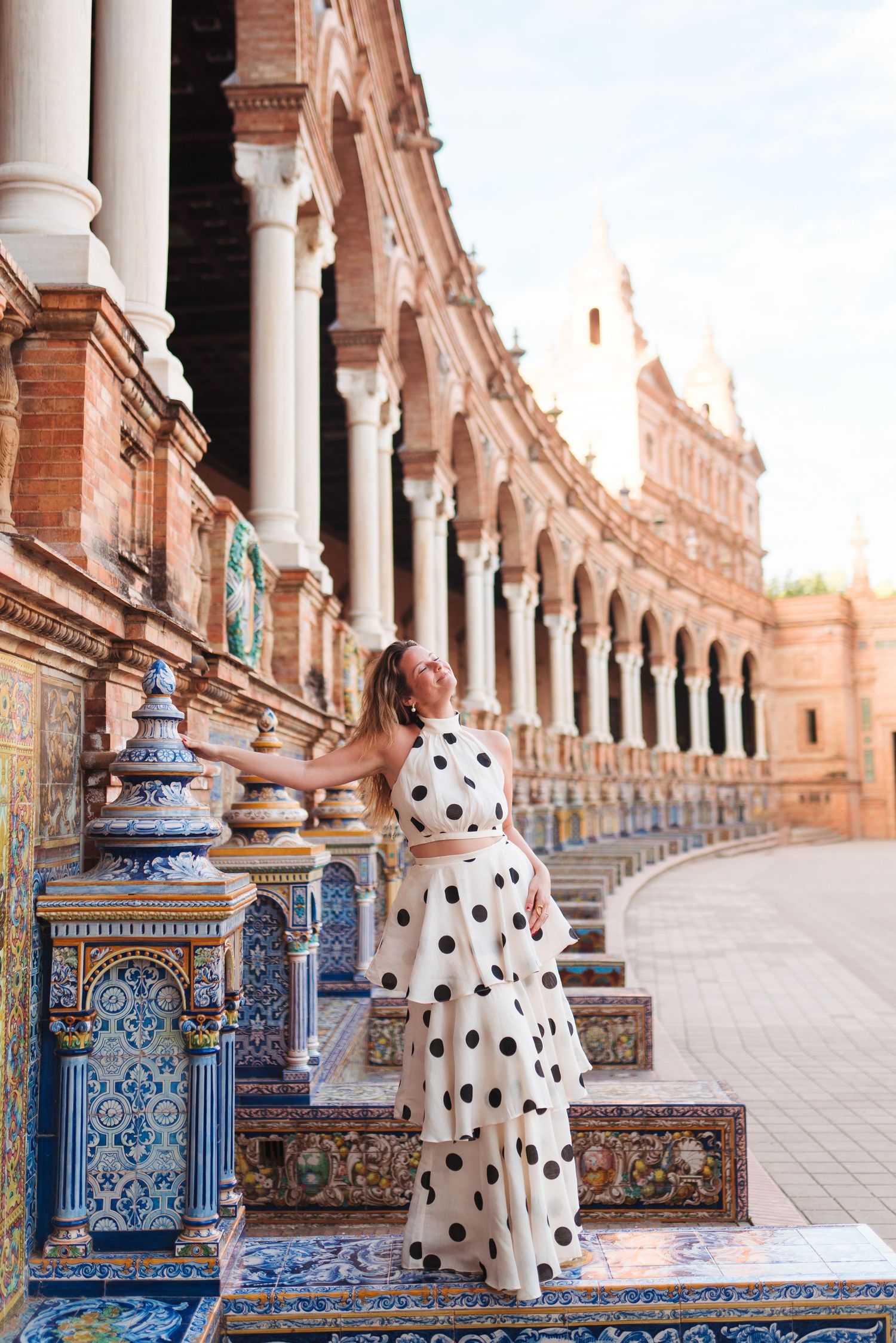 Woman in polka dot dress posing at Plaza de España, Seville, with ornate tiled benches and arched colonnade.