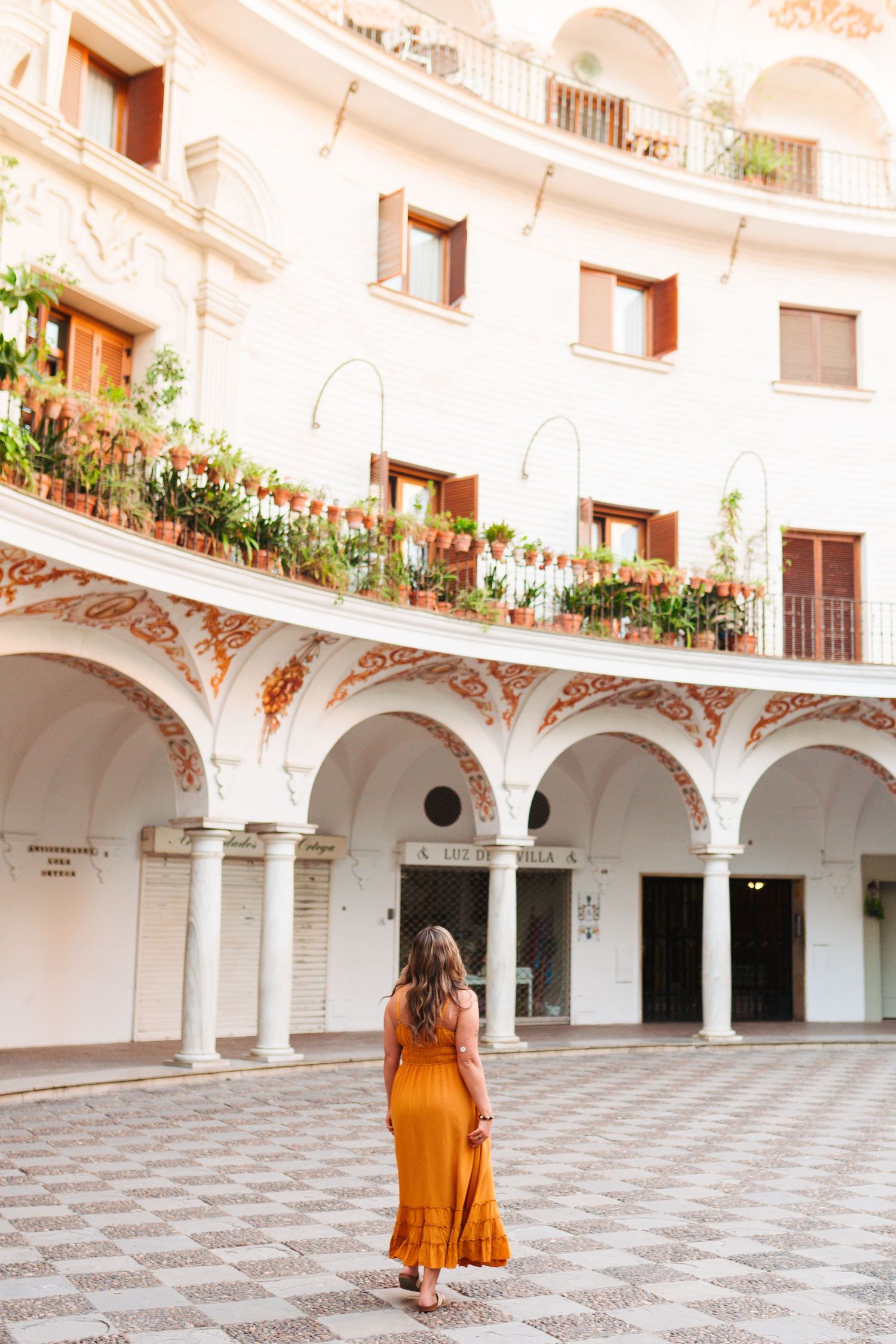 Woman in orange dress standing in elegant white arched courtyard with ornate details and flower-lined balconies.