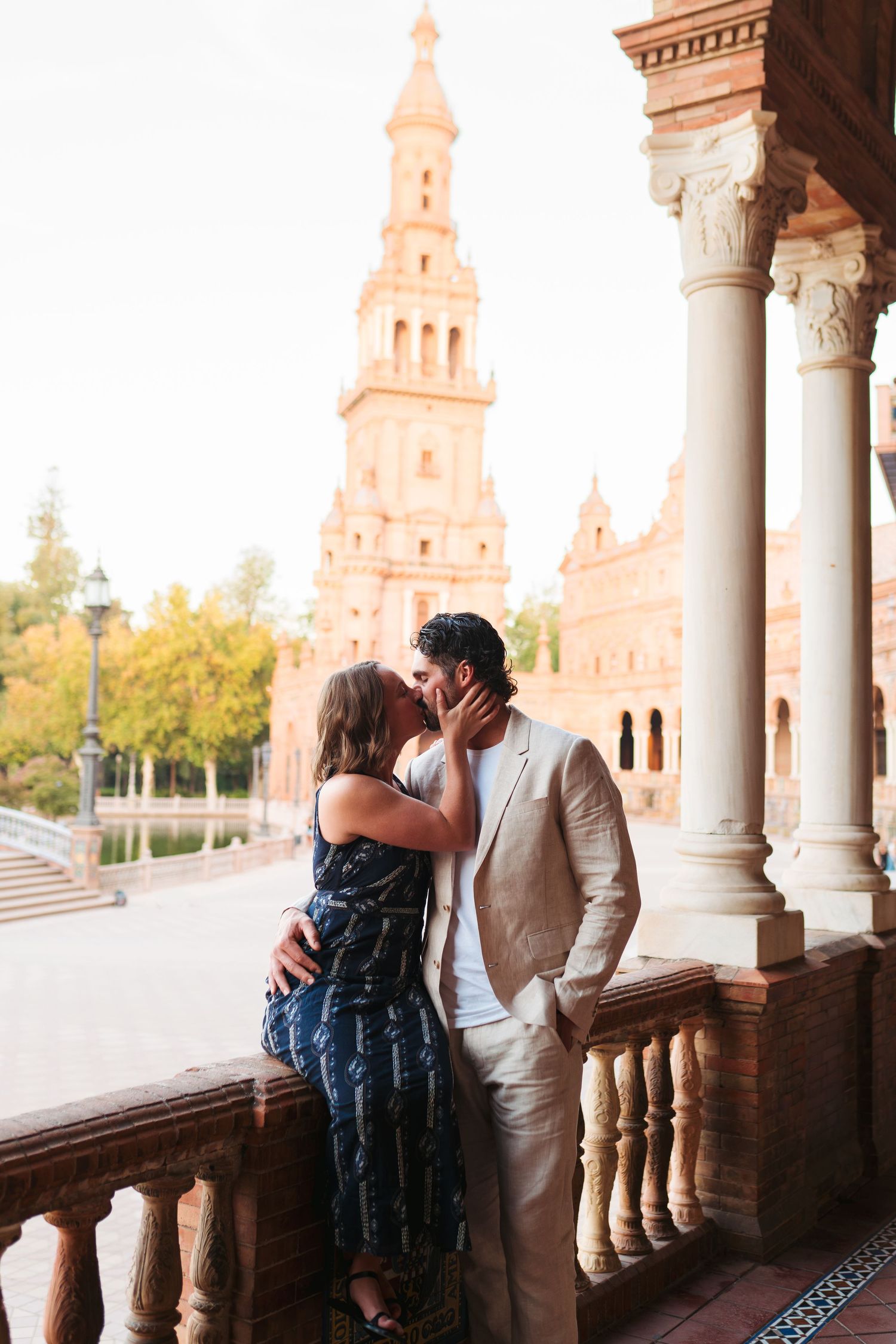 Couple kissing on a balcony at Plaza de España in Seville, Spain, with ornate tower and columns in background.